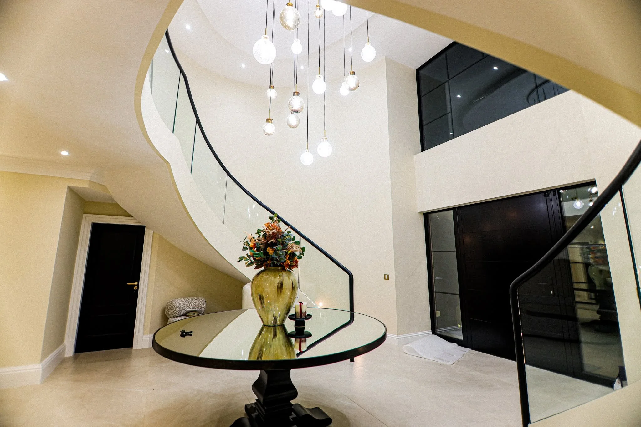 Interior of a modern house with a curved staircase, black handrails, pendant lights, a round table with a flower arrangement and candles, beige walls, and black doors and windows.