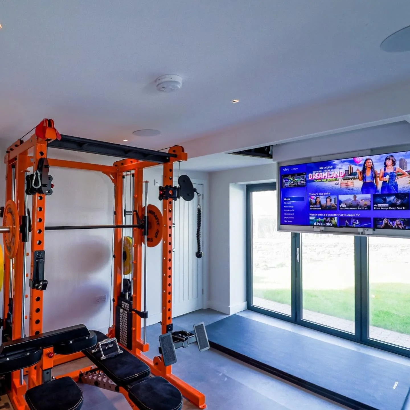 Home gym with orange weightlifting equipment, including a squat rack and weights, positioned near a large window and a wall-mounted TV displaying streaming content.