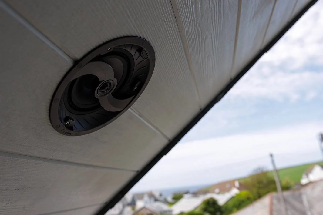 Outdoor speaker mounted under roof edge with a view of houses, trees, and overcast sky in the background.