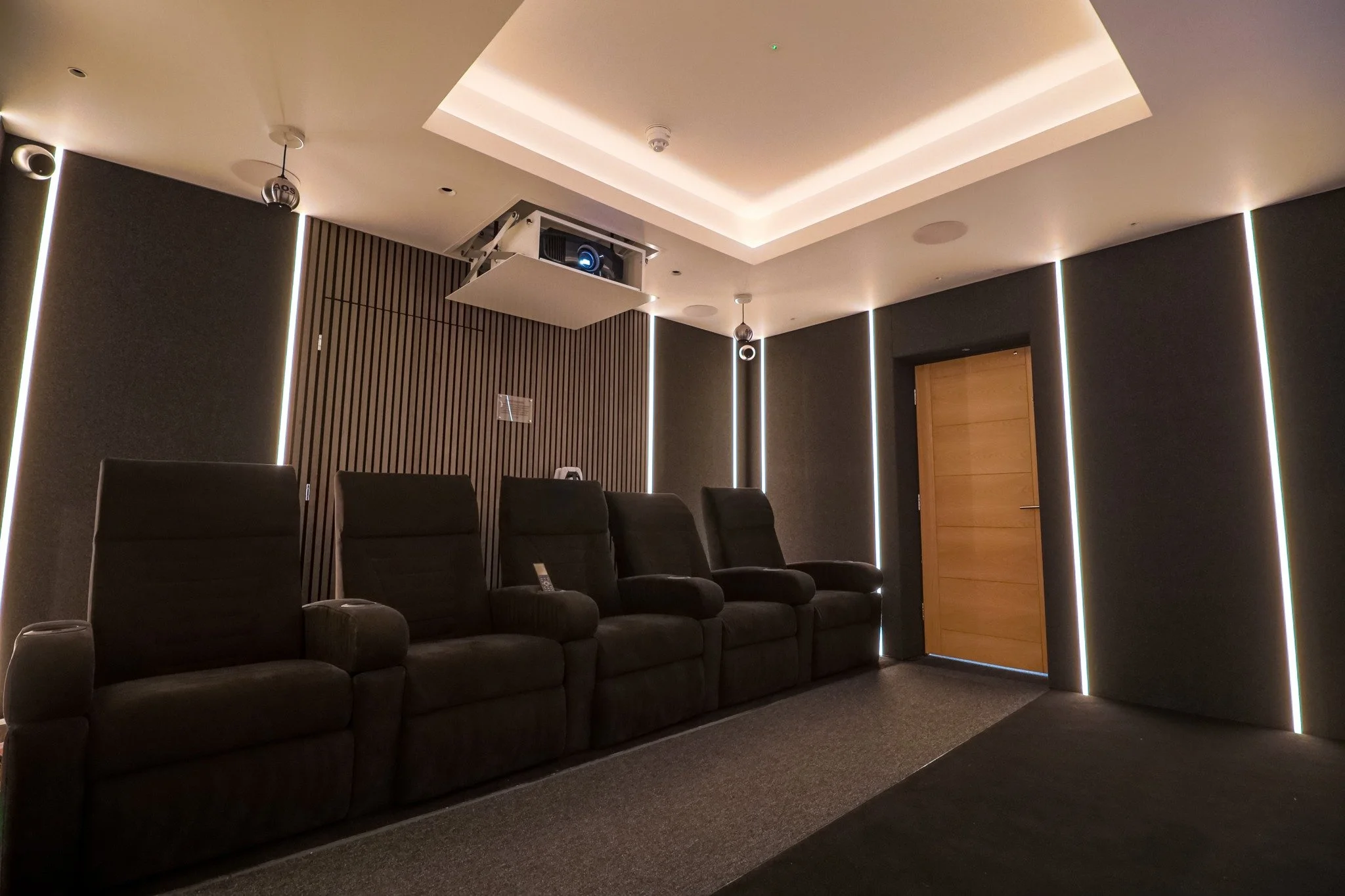 A modern, dark-themed home theater room with four black recliner chairs, wooden paneling on the wall, and vertical LED lighting strips. There is a ceiling-mounted projector and a wooden door on the right side.