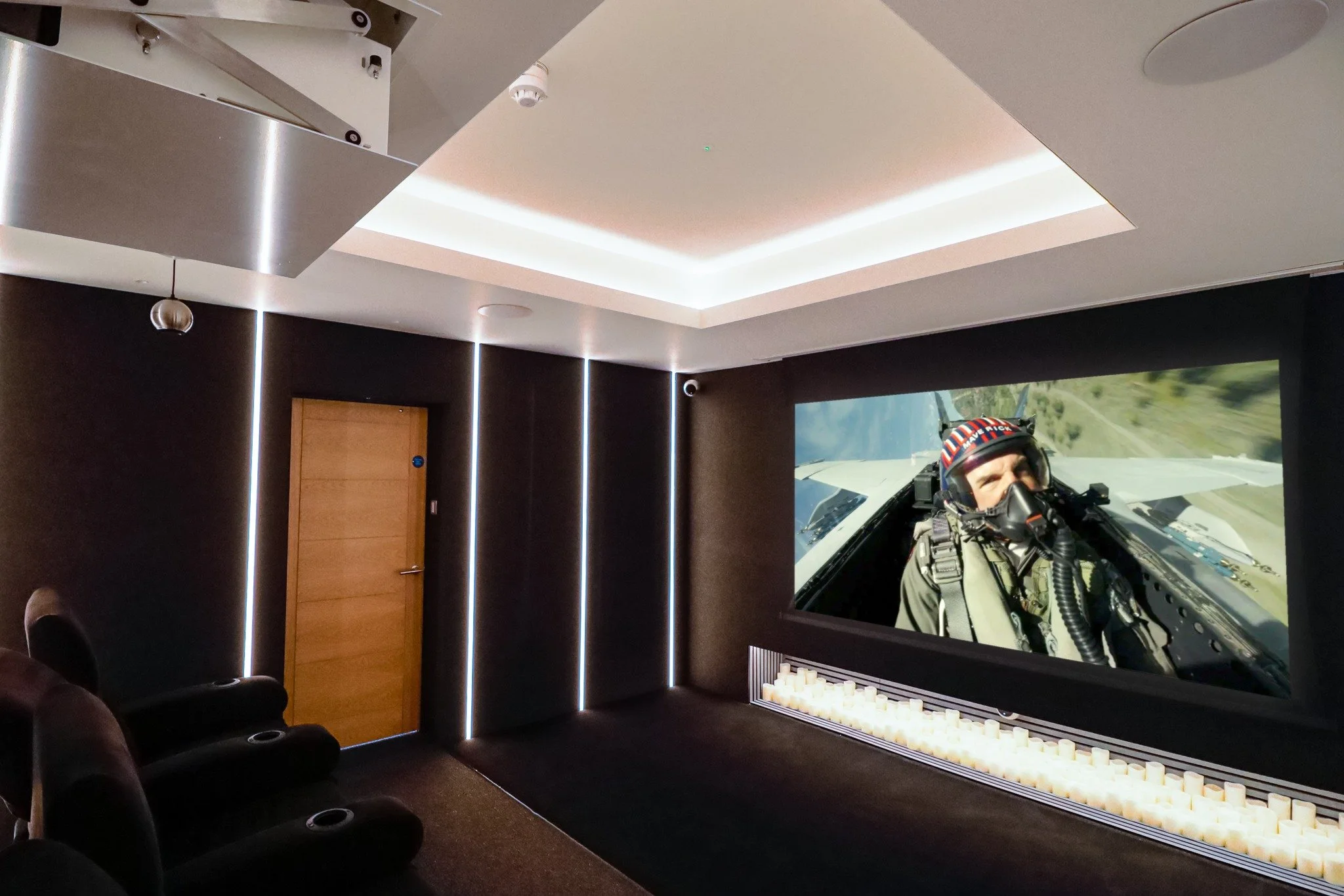 Home theater room with dark walls, a large screen showing a fighter pilot, and black reclining chairs.