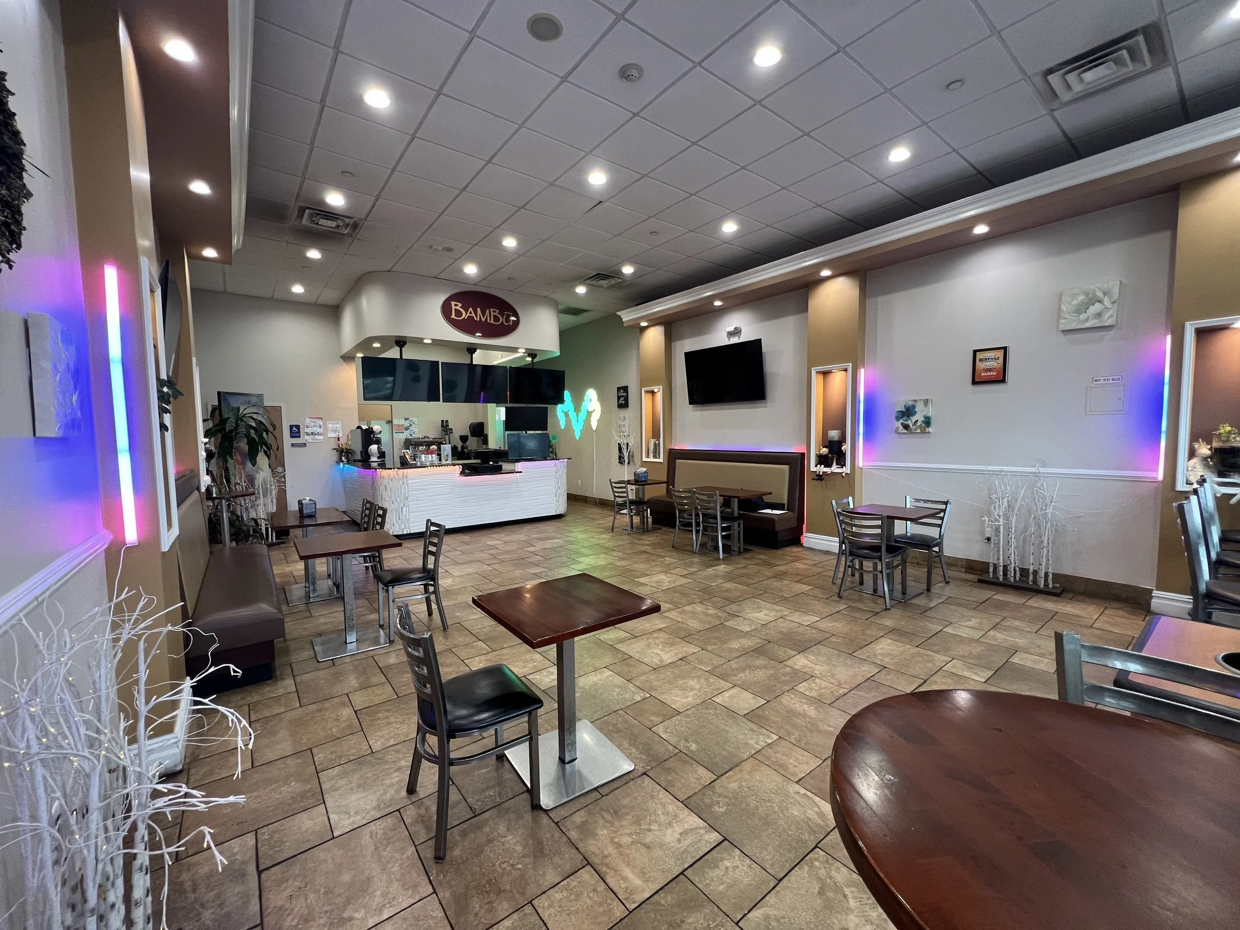 Interior of a casual restaurant or cafe with tables and chairs, a counter area with a sign that says 'Bambu,' and colorful neon lights along the walls.