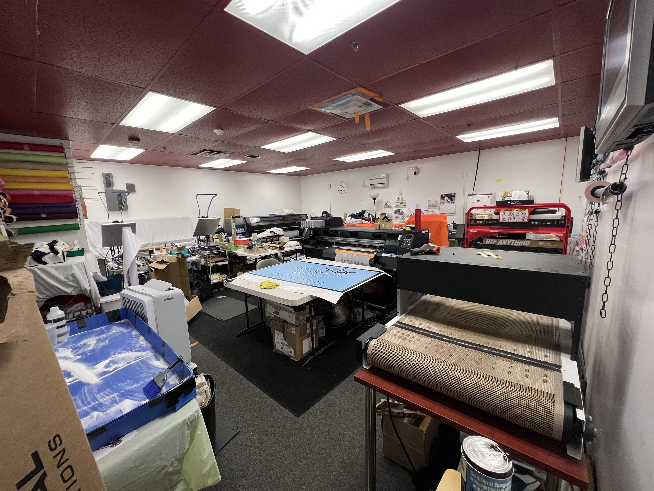 A cluttered print shop or sign-making workspace with printers, tables, a large cutting or plotting machine, and various supplies scattered around.