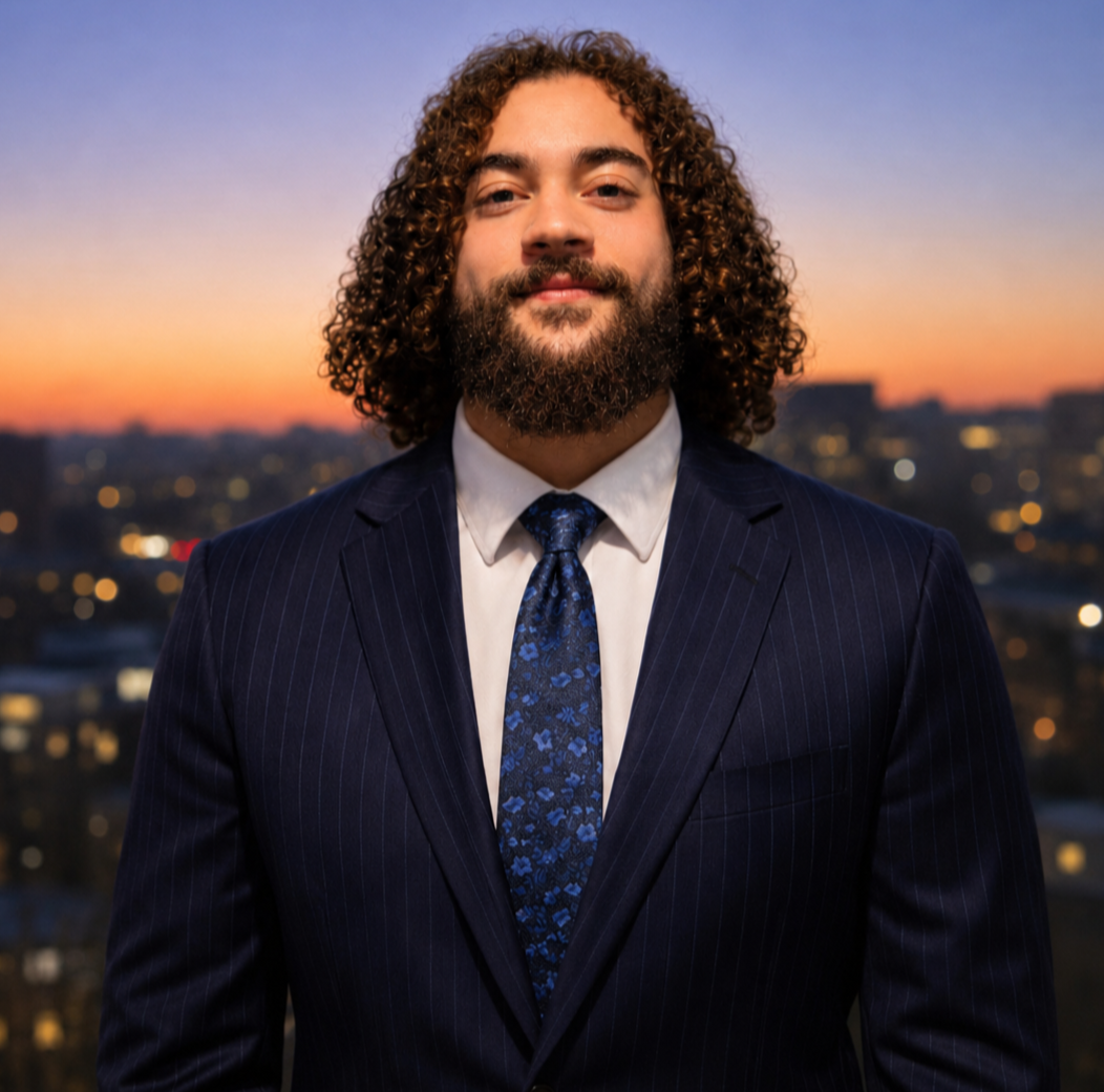 A man with curly hair and a beard in a dark blue pinstripe suit, white shirt, and patterned tie standing against a sunset cityscape background.