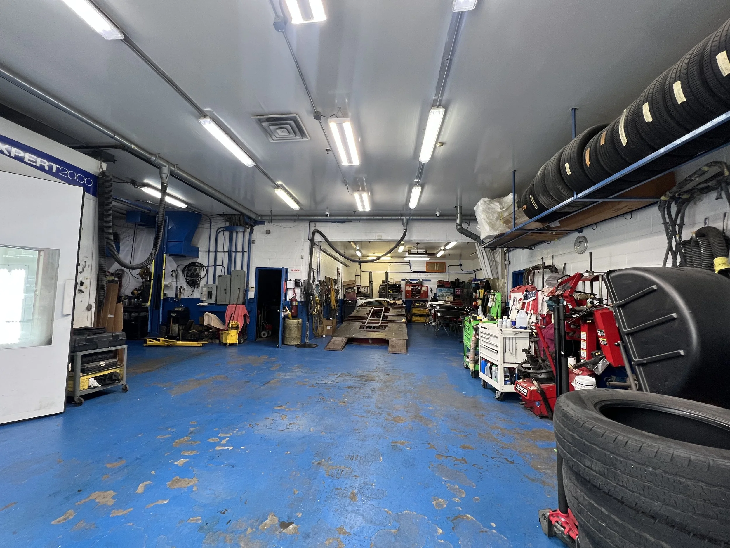 A well-organized automotive repair shop with blue painted concrete floor, fluorescent ceiling lights, and various tools, tires, and equipment stored along the walls and on shelves.