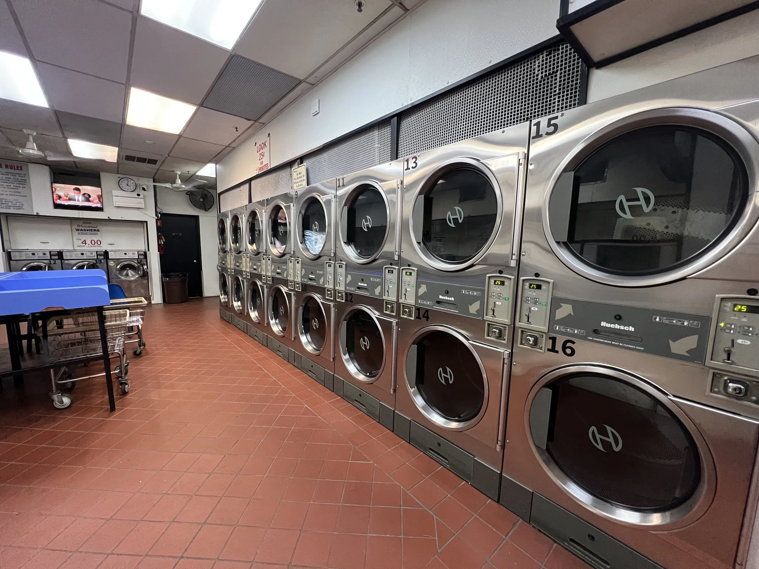 Laundromat with multiple industrial washing machines and dryers, coin-operated, with a few blue baskets and a small TV on the wall.