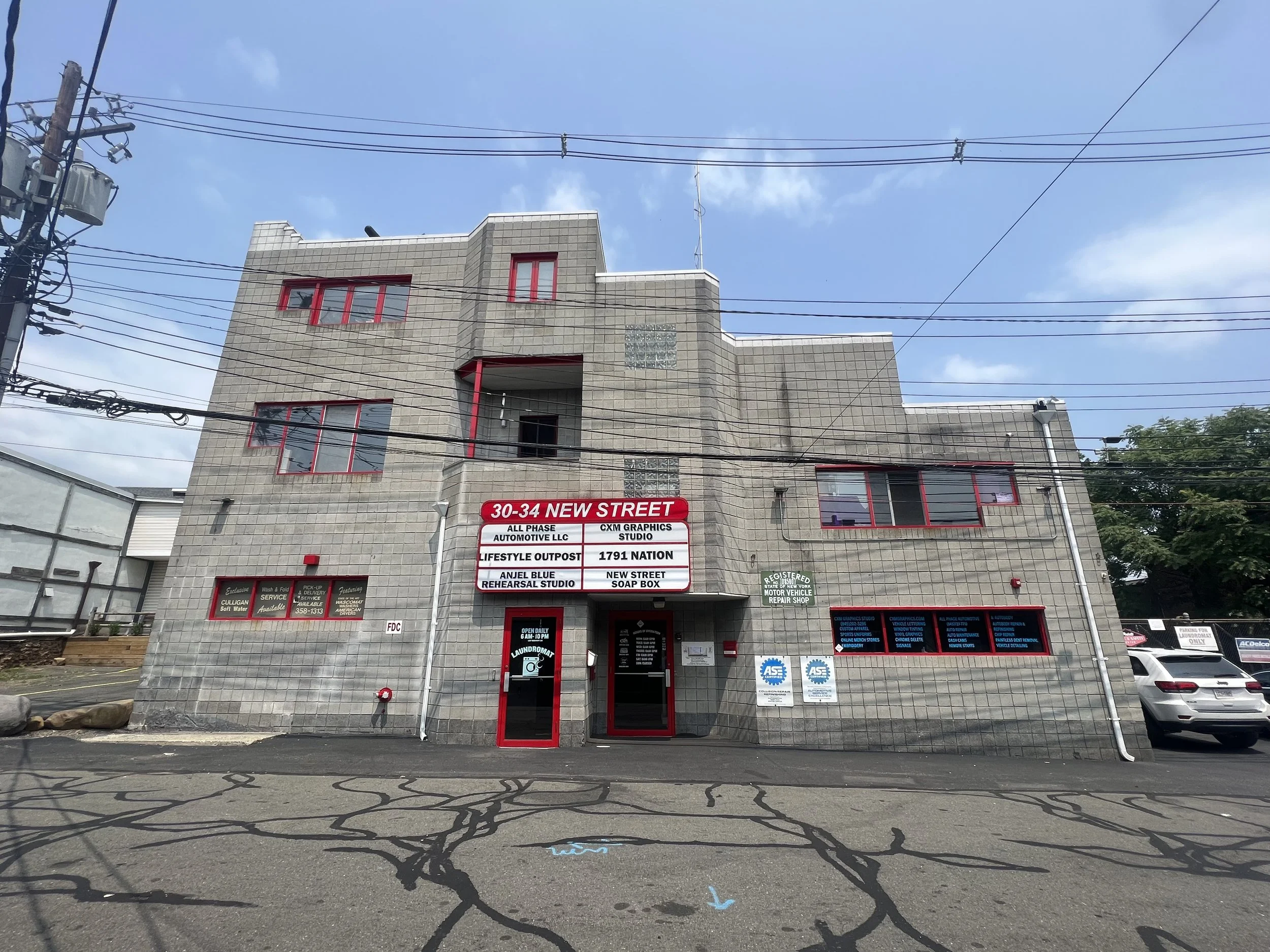 A multi-story building with gray brick exterior and red window frames. A sign on the building displays various business names and addresses. The building is located on a paved street with electrical wires overhead and some parked cars on the right. The sky is partly cloudy.