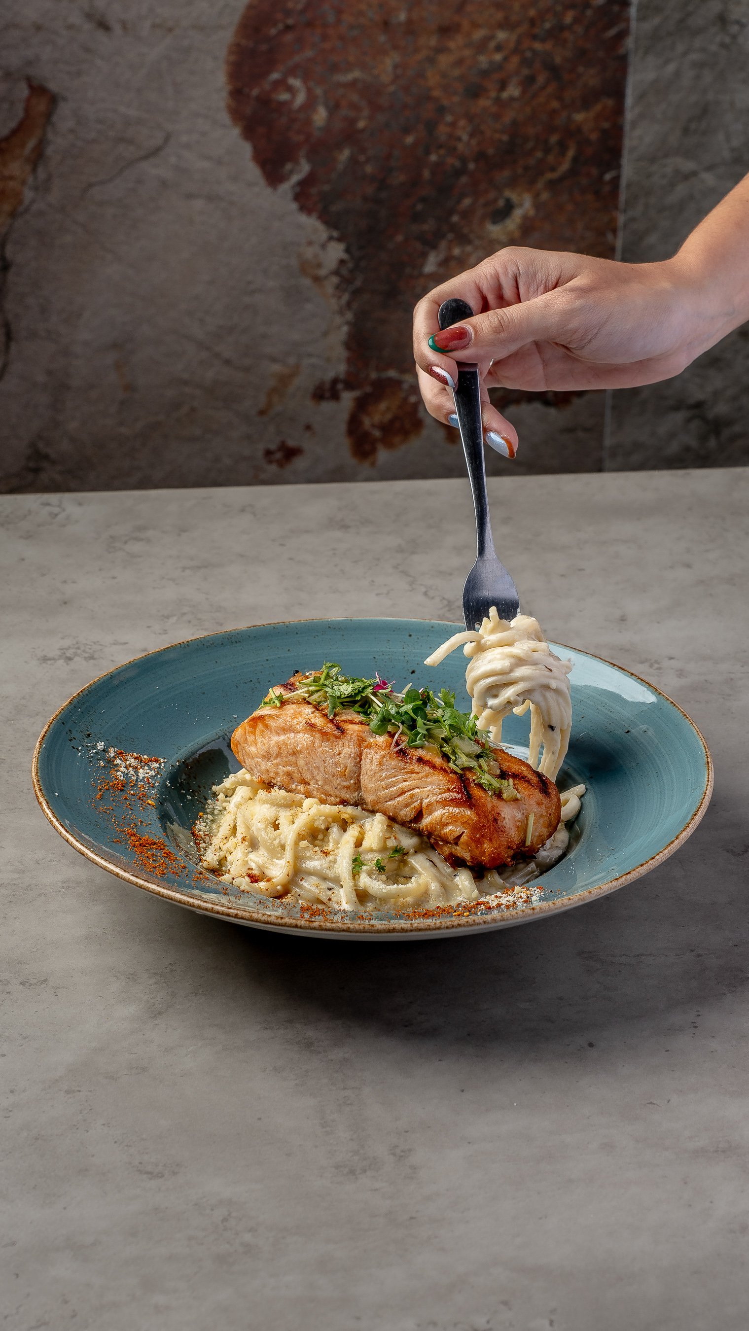 A plate of fettuccine pasta topped with grilled salmon, diced vegetables, and fresh herbs, with a hand holding a fork above the dish.