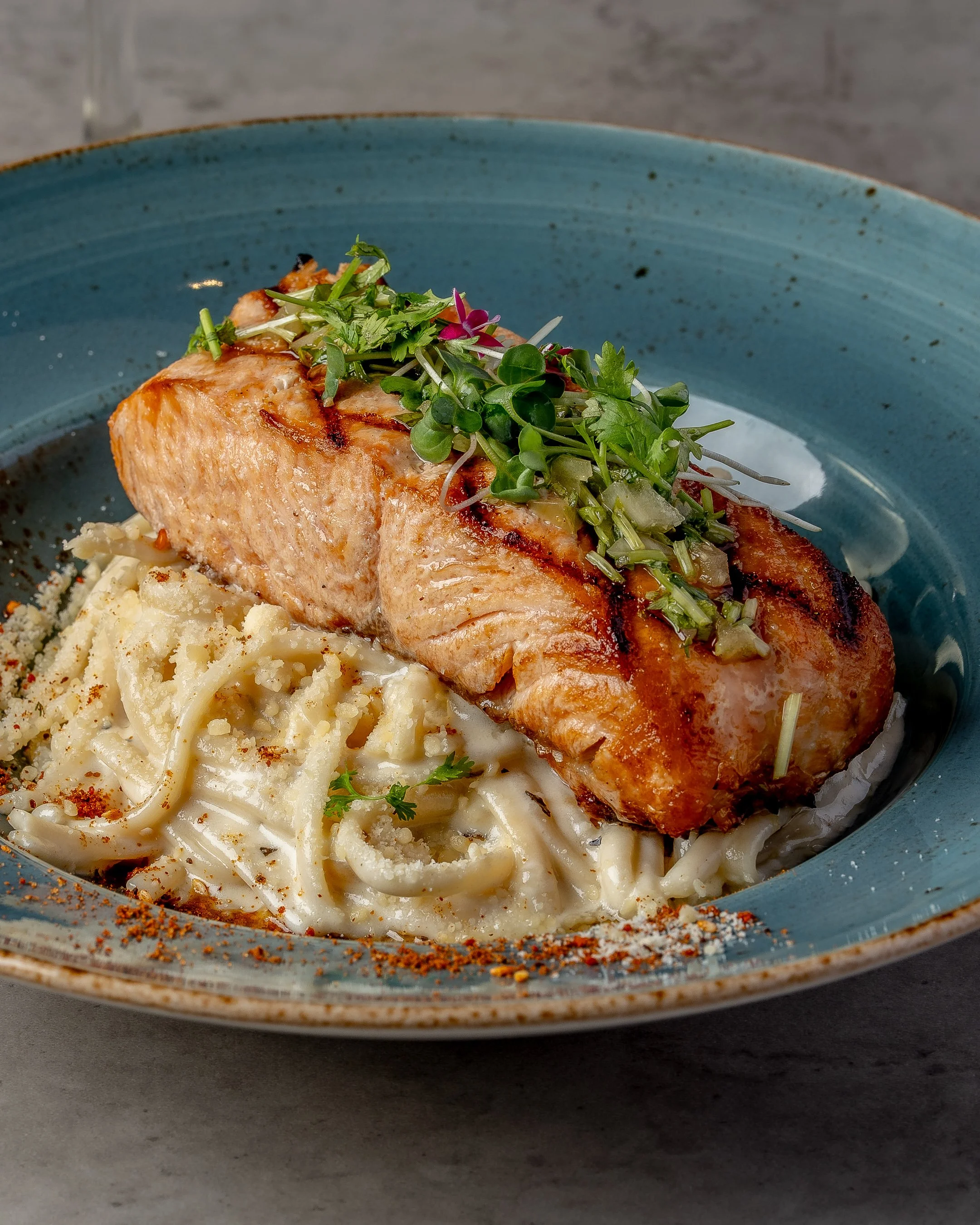 A plate of cooked salmon topped with microgreens and served on a bed of creamy pasta, garnished with spices.