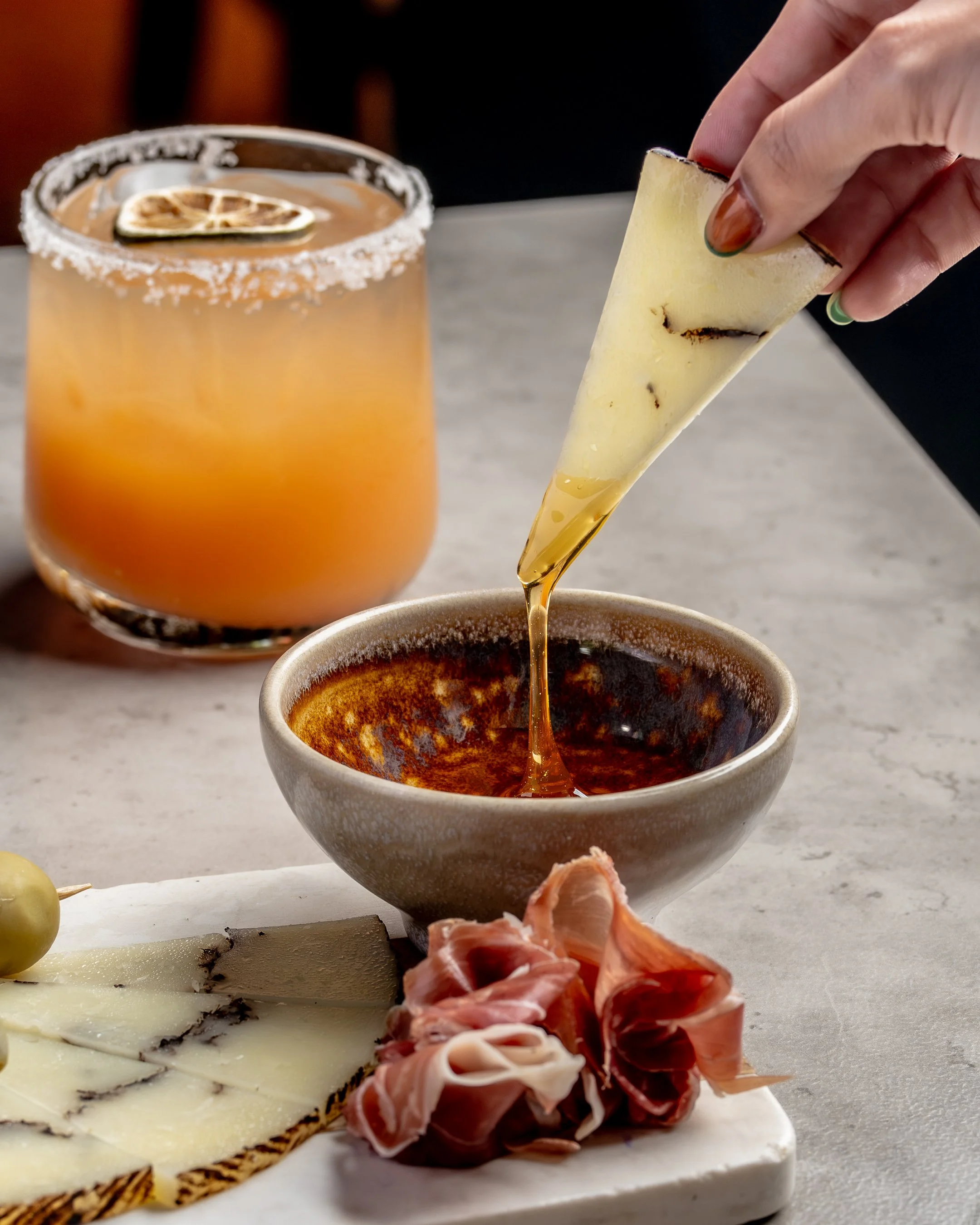 A person pouring honey into a bowl with charcuterie and cheese on a marble board, a glass of orange cocktail with a lemon slice on the rim in the background.