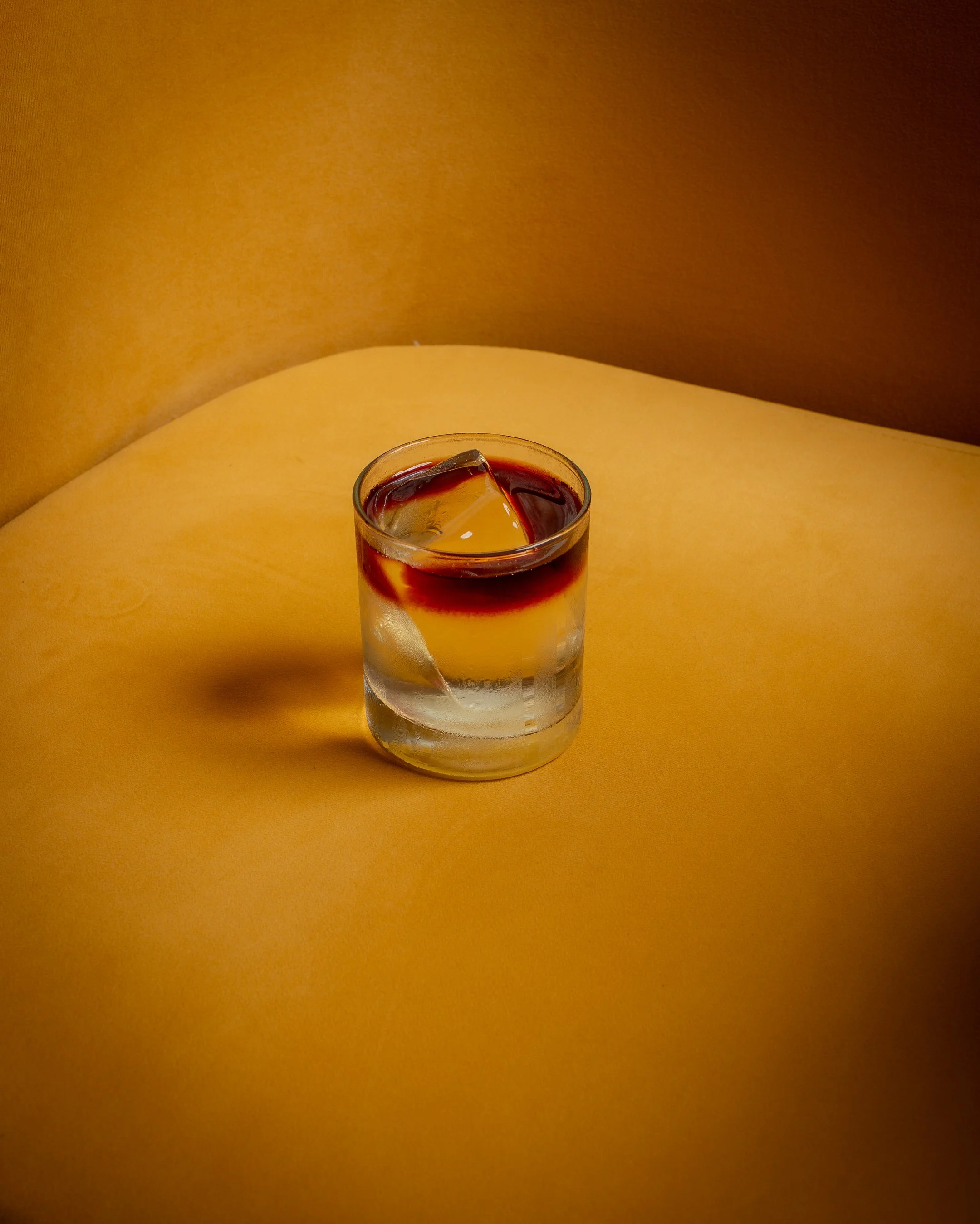 A clear glass filled with a dark red beverage, with ice cubes inside, placed on a yellow surface against a brown background.