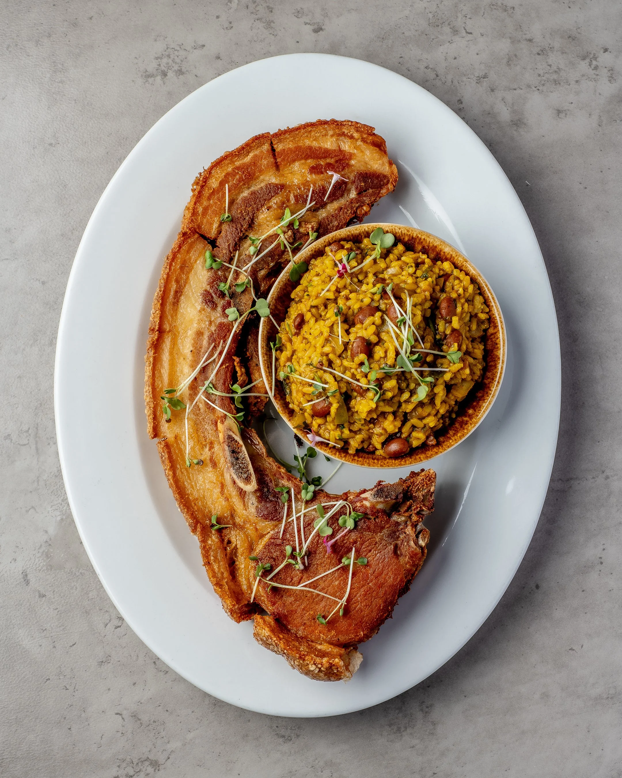 A white oval plate with two slices of crispy pork belly and a small bowl of yellow rice with beans and herbs, garnished with microgreens, on a gray textured surface.