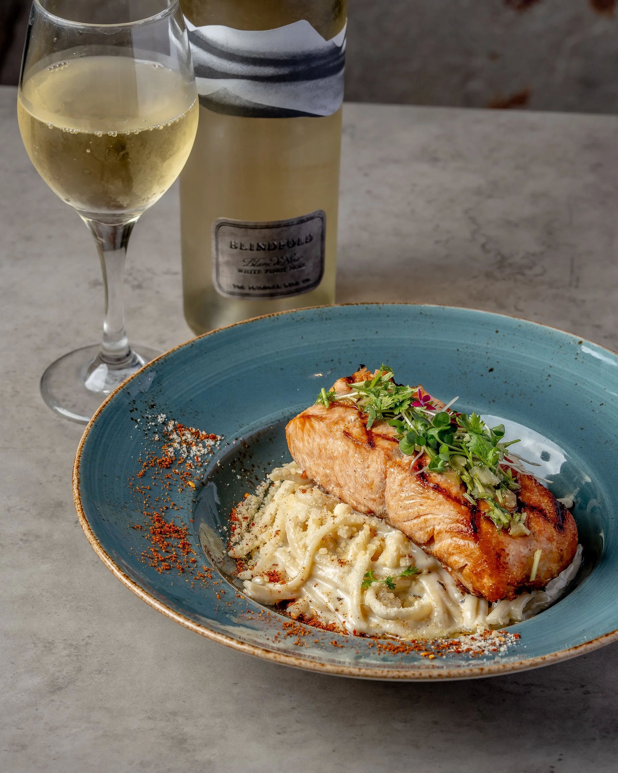 A plate of creamy pasta with grilled salmon and fresh greens, garnished with spices. A glass of white wine and a bottle of wine are in the background on a gray table.