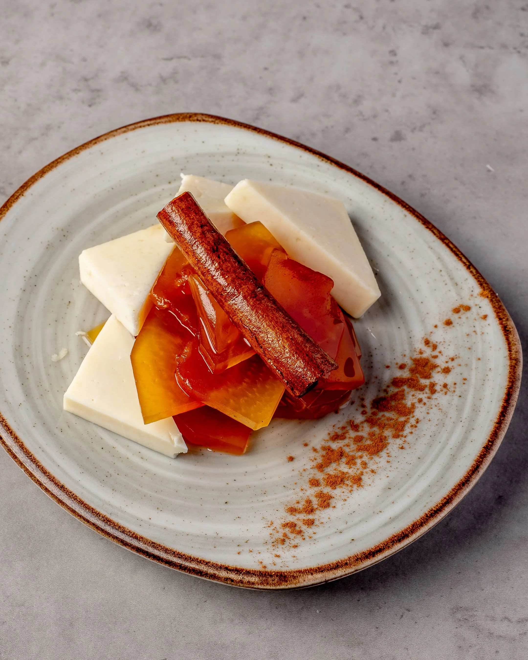 Close-up of a ceramic plate with slices of cheese, a slice of yellow bell pepper, a tomato, and a cinnamon stick, on a gray surface.