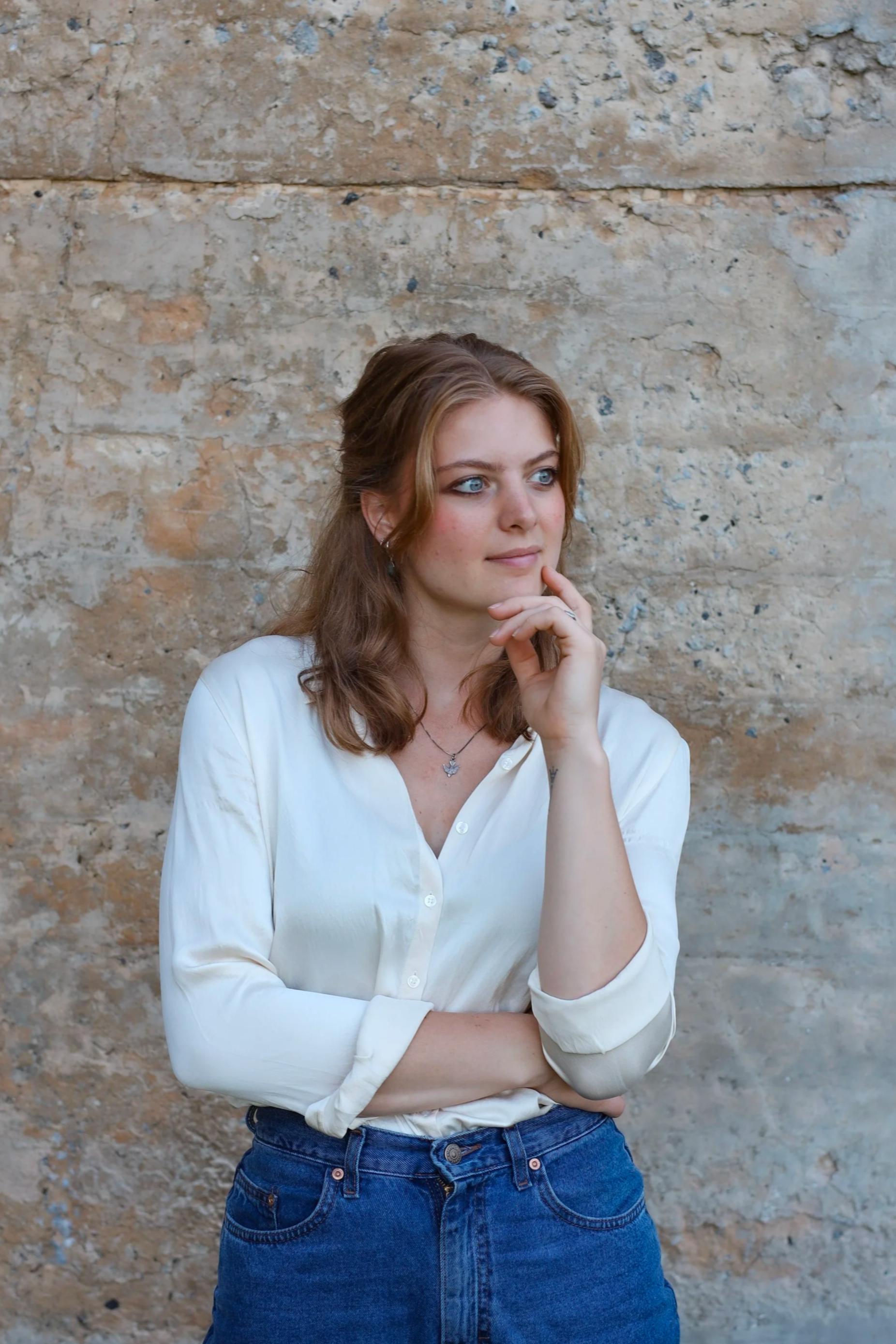 A young woman with wavy brown hair, blue eyes, wearing a white button-up shirt and blue jeans, standing against a textured stone wall. She has her left arm crossed and her right hand resting on her chin, appearing thoughtful.