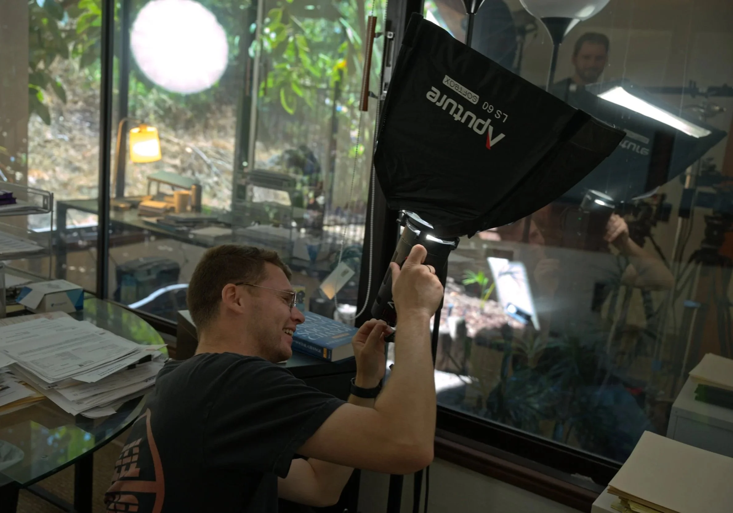 A man smiling and wearing glasses, holding a photography umbrella inside an office with a glass window, books, papers, and a reflection of another person in the background.