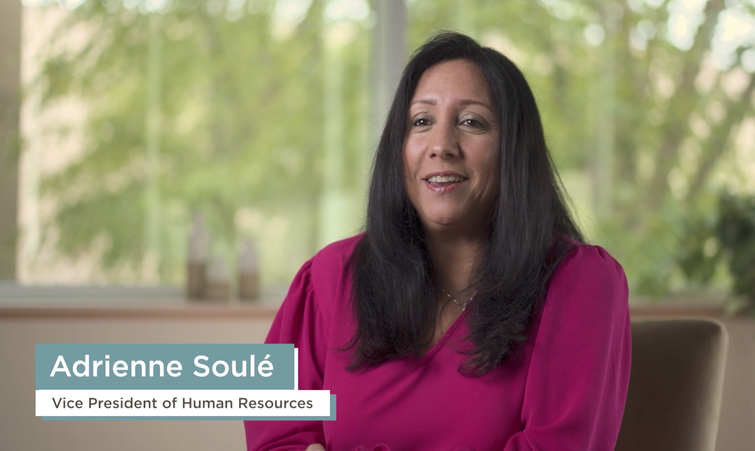 A woman with black hair wearing a pink blouse, sitting indoors in front of a large window with green trees outside, smiling. The text overlay identifies her as Adrienne Soulé, Vice President of Human Resources.