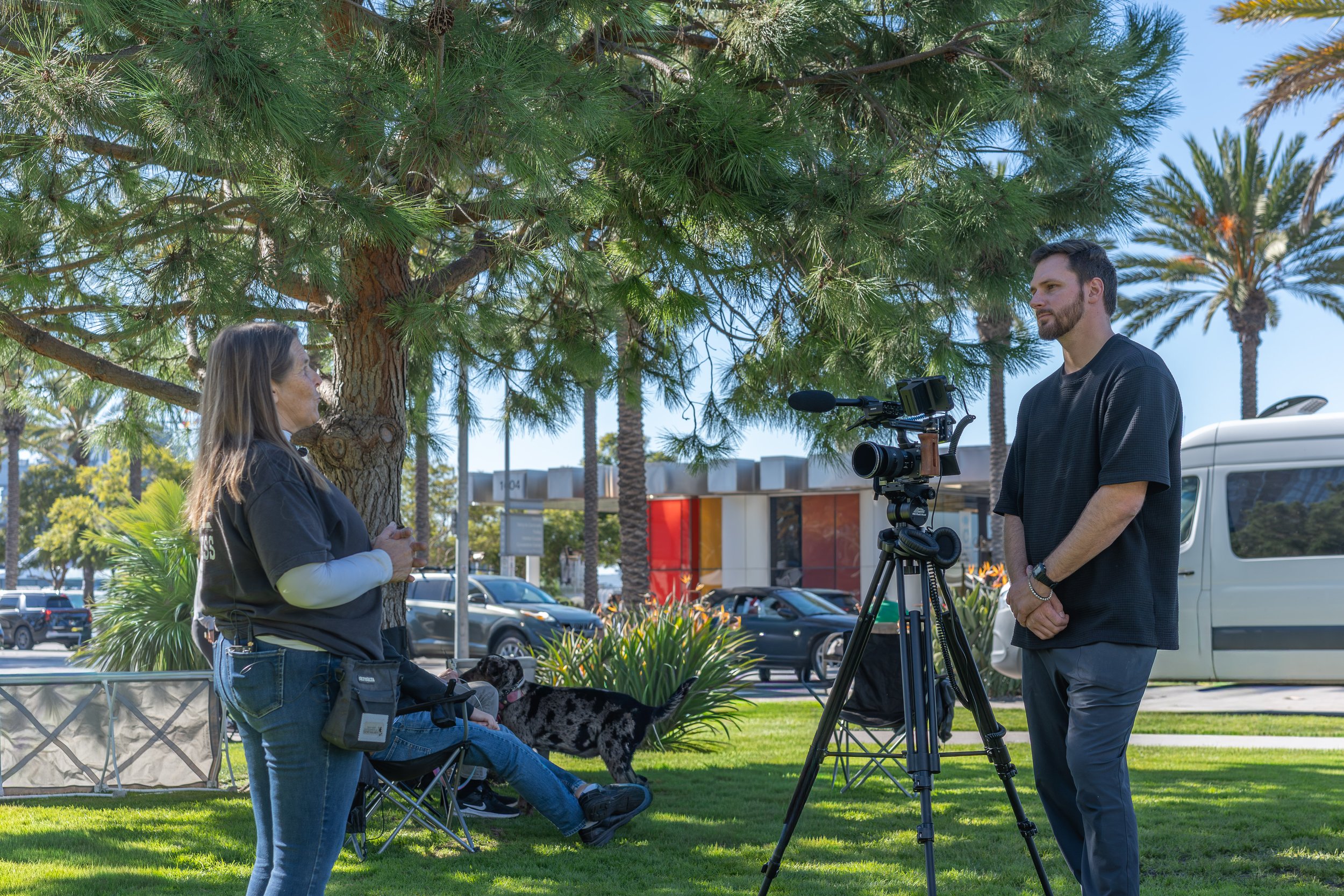 A woman being interviewed outdoors on a sunny day, standing near a tree, while a man with a camera records her. There are cars and palm trees in the background.