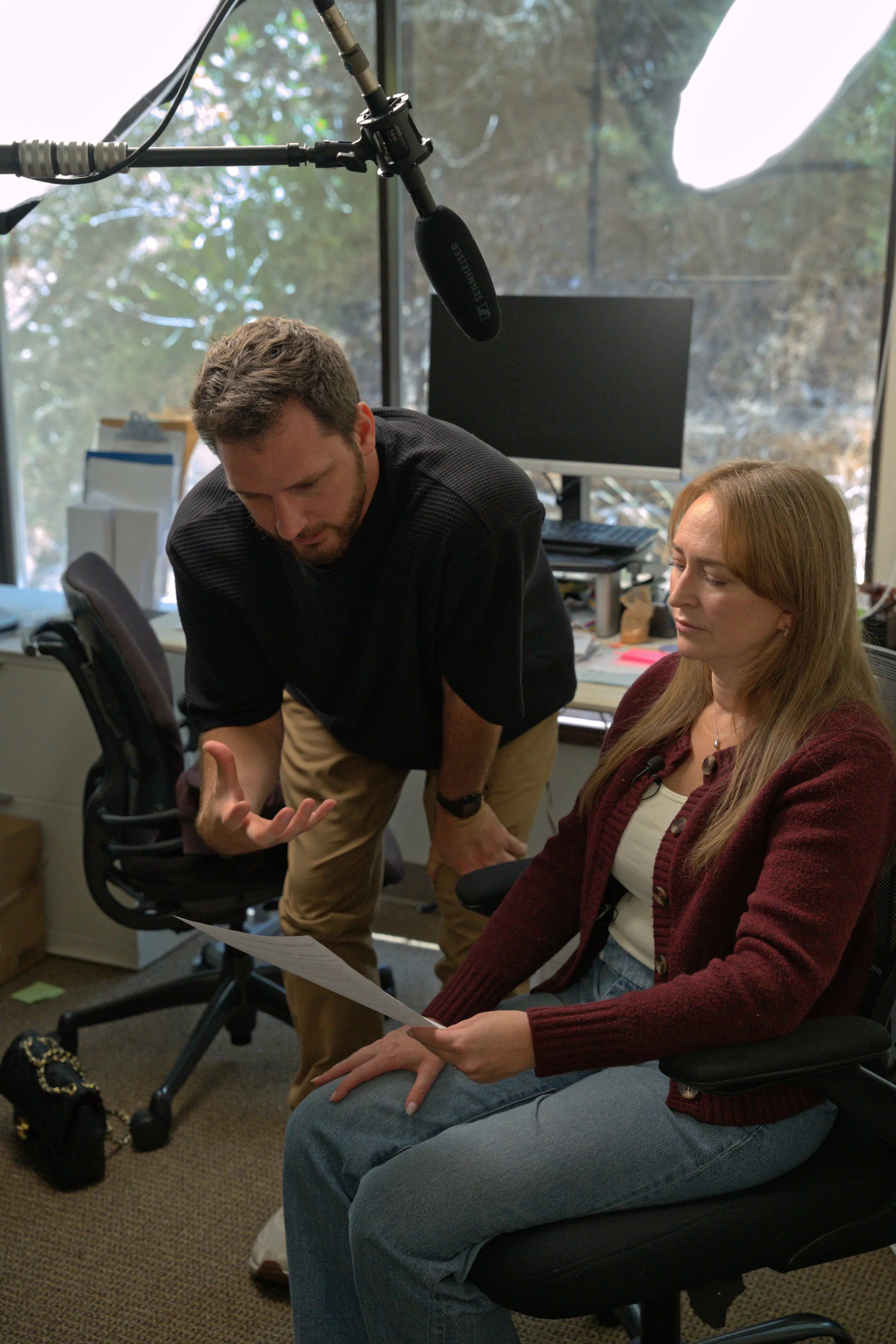 A man with brown hair and beard showing a document or paper to a woman with blonde hair, who is sitting on a black office chair in an office with large windows and a computer monitor in the background. The woman is wearing a maroon sweater and jeans, and is sitting on her lap. The man is leaning over and gesturing with his hand. There is a boom microphone hanging from above.