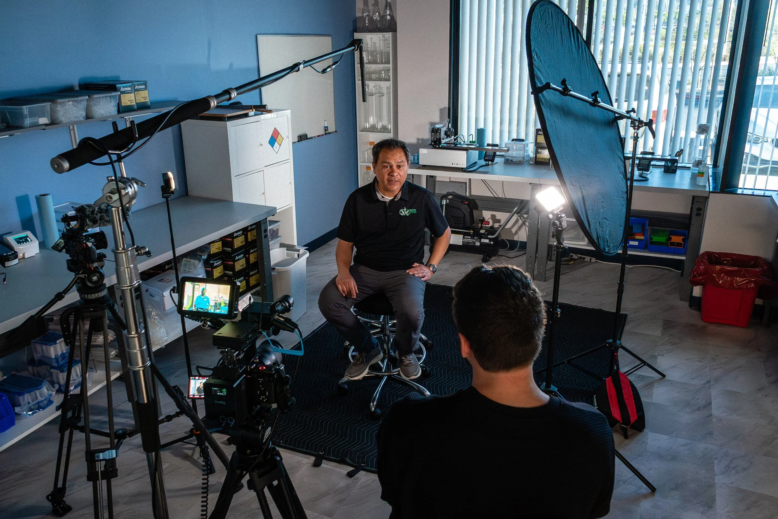 A man being interviewed or recorded in a professional setting with cameras, lighting, and equipment, sitting in front of a blue wall with shelves and a window with vertical blinds nearby. Filmed by a San Diego video production company.