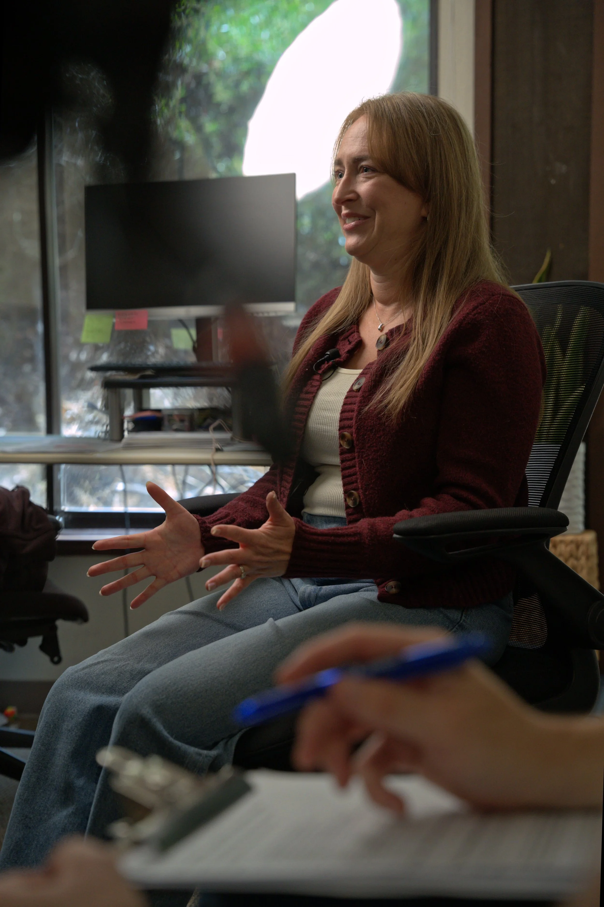 A woman with long, reddish-brown hair wearing a maroon cardigan and a light-colored shirt is sitting in an office chair during a discussion. She appears to be smiling and gesturing with her hands. There is a computer monitor, some documents, and a window with green foliage outside in the background.