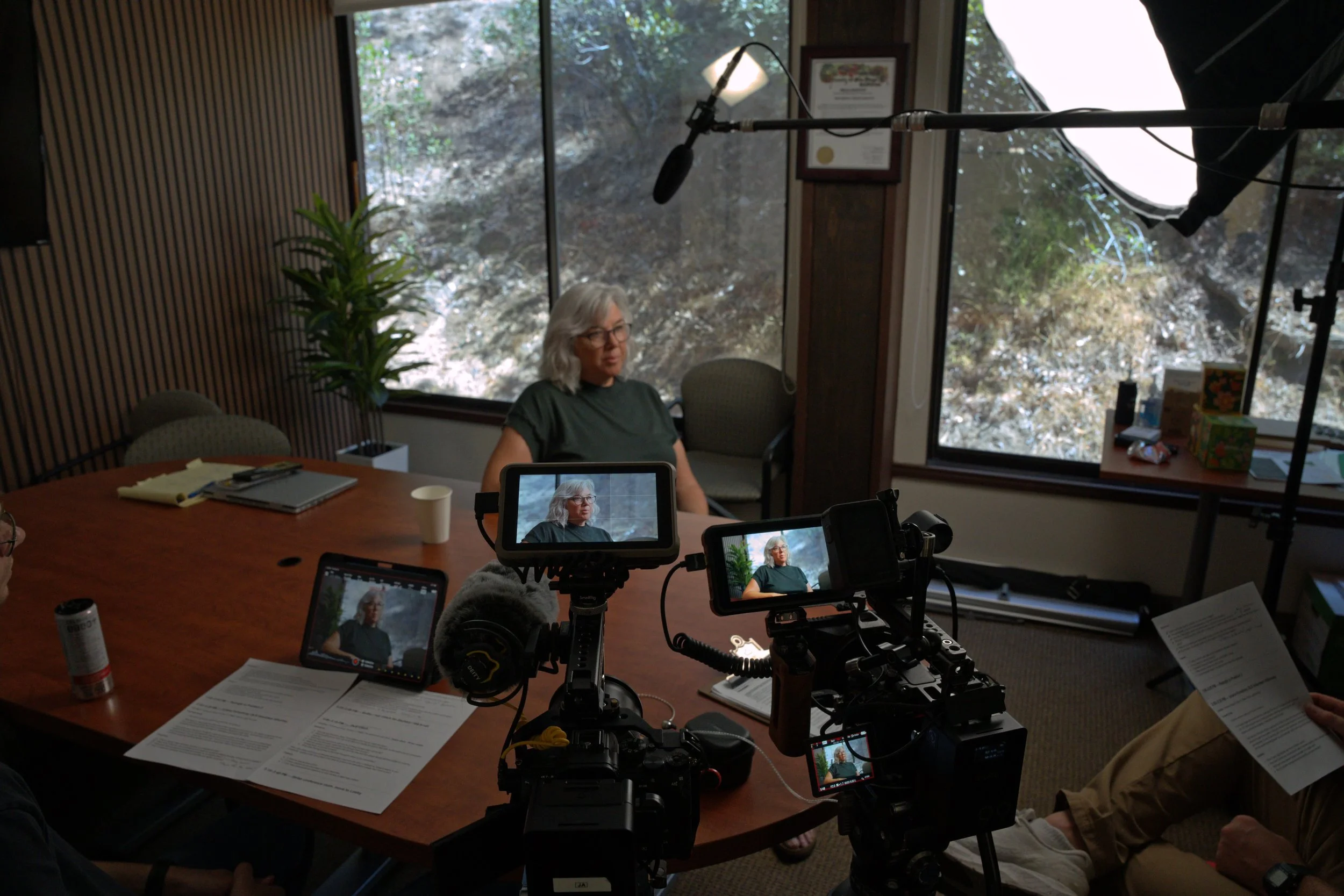 A woman with gray hair and glasses is sitting at a conference table during a video recording or interview session. Multiple cameras and filming devices are capturing her, and a large window behind her shows trees outside. The room has wood-paneled walls and a few items like a plant, documents, and a coffee cup on the table.