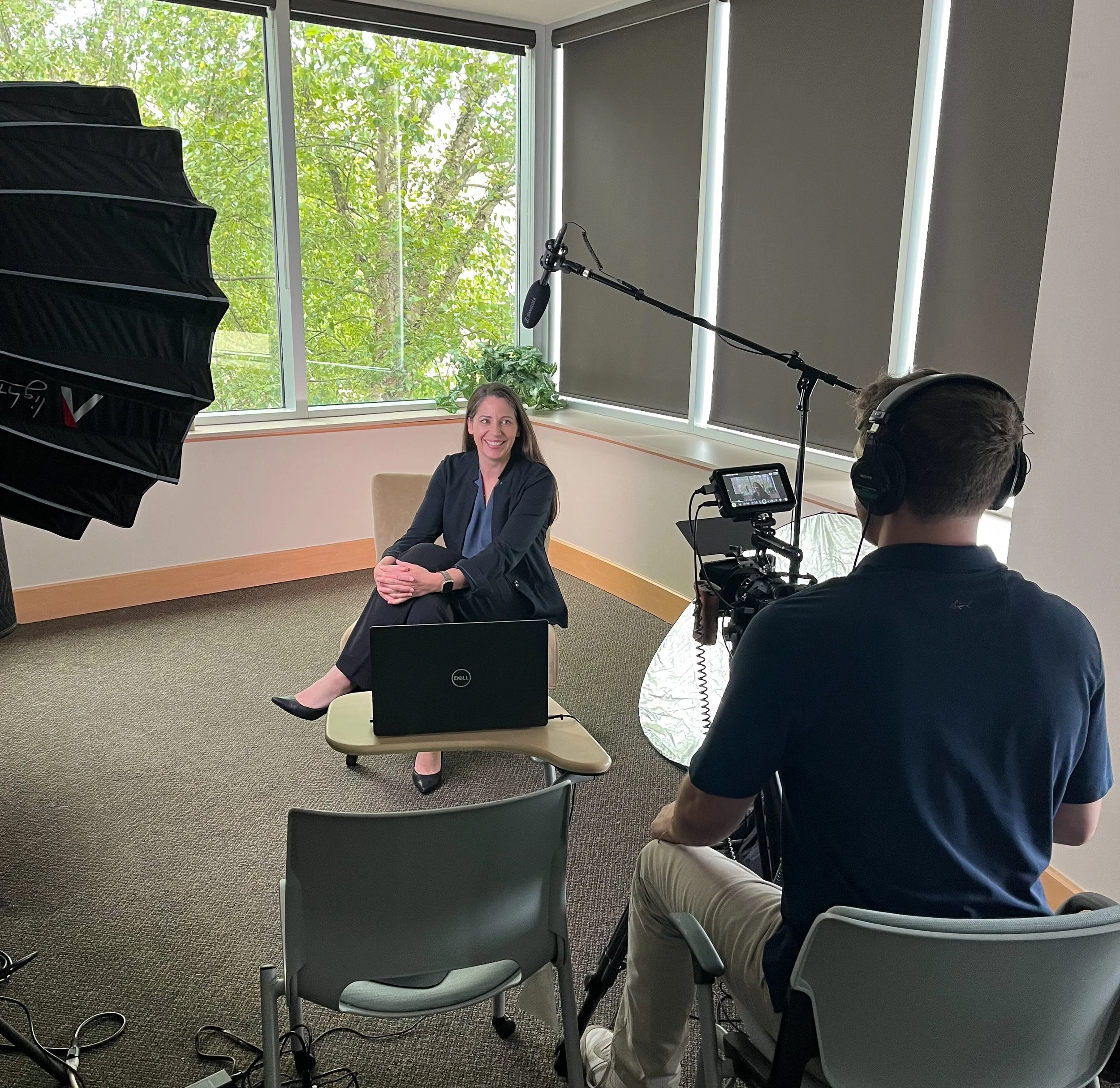 A woman in a dark blazer sitting on a beige chair, being filmed by a man using a professional camera in an office setting. The setup includes a large light on the left, a laptop on a small table, and a window with green trees outside.