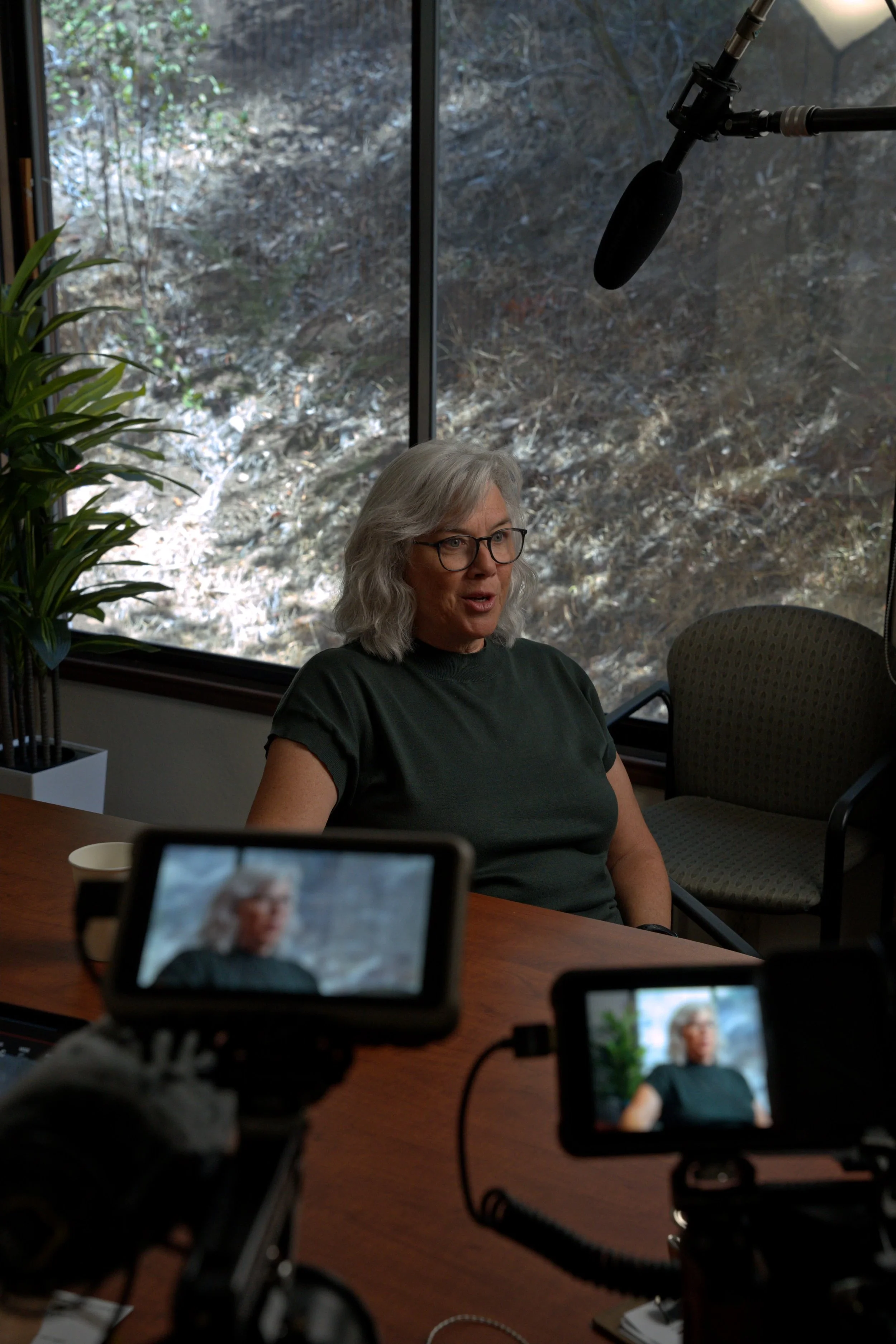 A woman with gray hair and glasses is sitting at a table during an interview or conversation, with multiple cameras and microphones capturing her from various angles. There is a window behind her revealing a rocky outdoor scene and a green plant to the left.