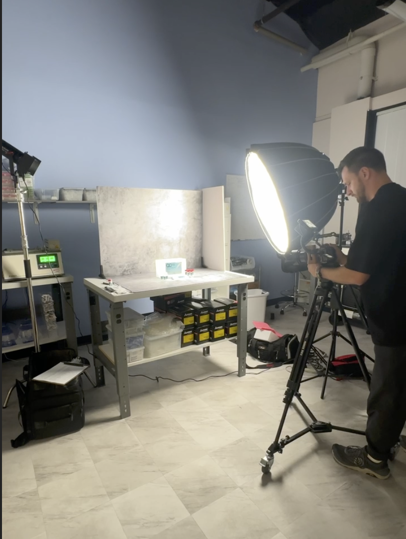 A man operating a camera with studio lighting aimed at a small digital clock on a work table, in a photography studio with equipment and storage boxes.