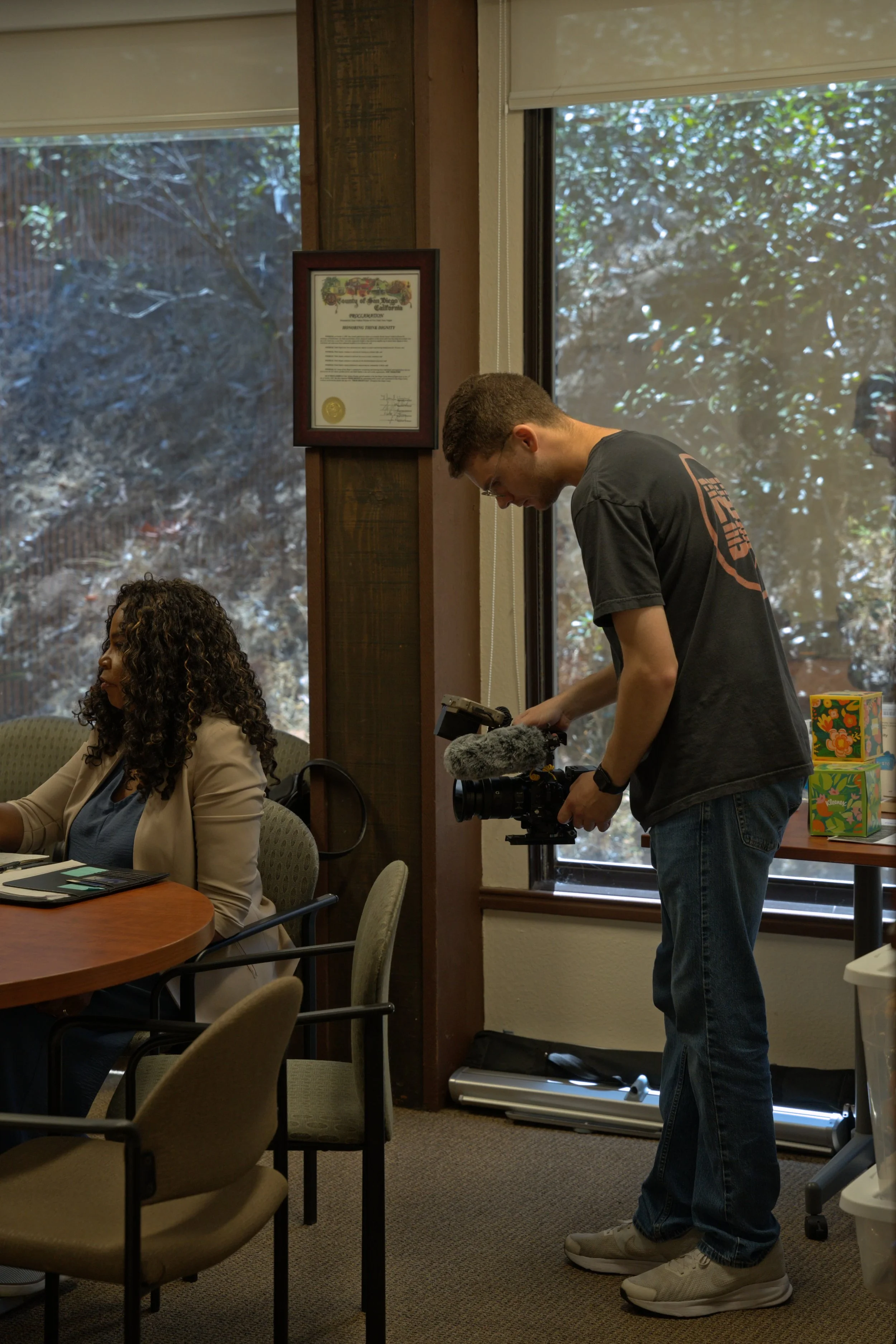 A man operating a camera on a tripod inside an office building with large windows and a framed document on the wall, while a woman is seated at a conference table.