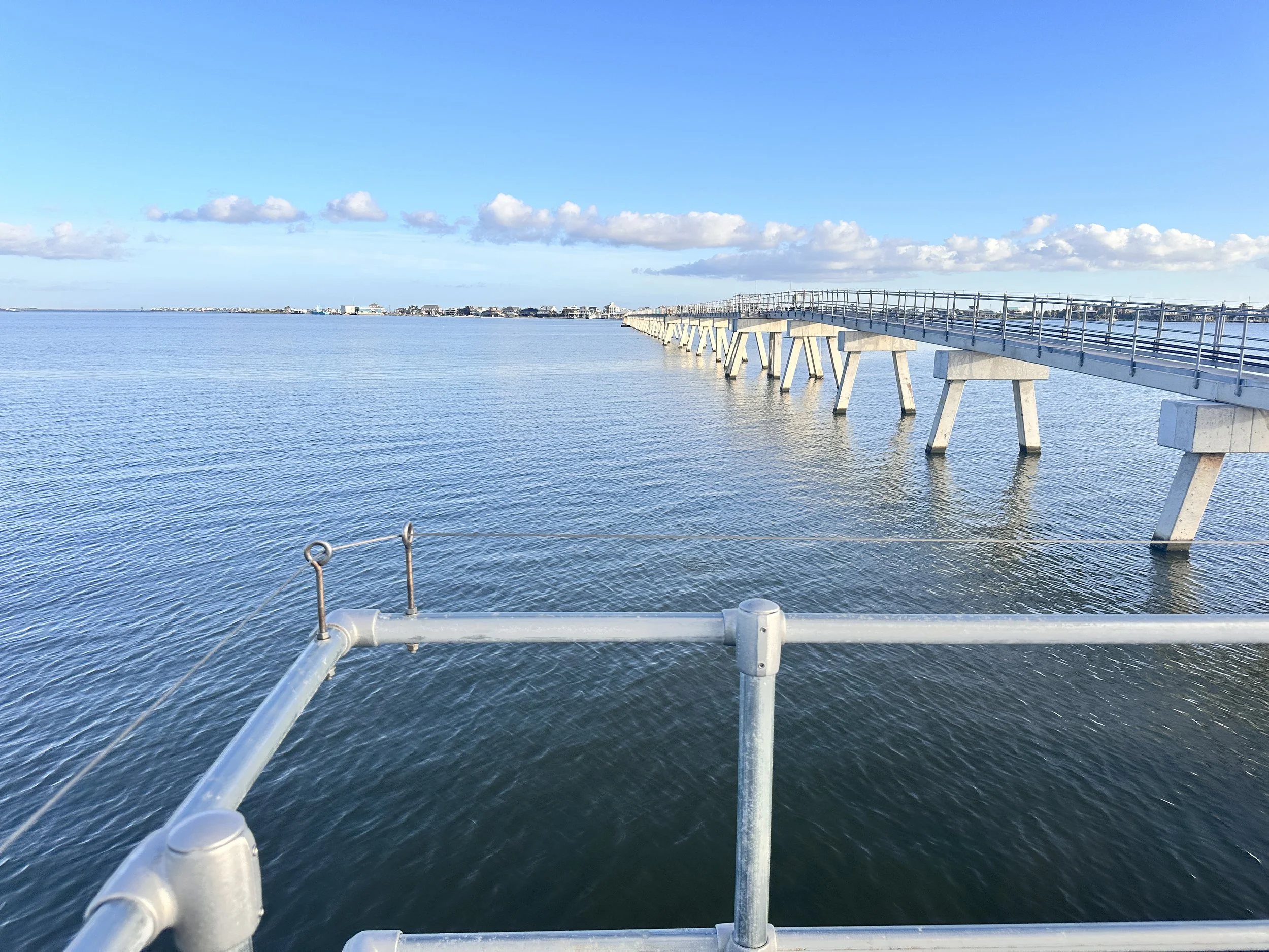 A pier extending over calm water with a railing in the foreground and a distant shoreline under a blue sky with scattered clouds.