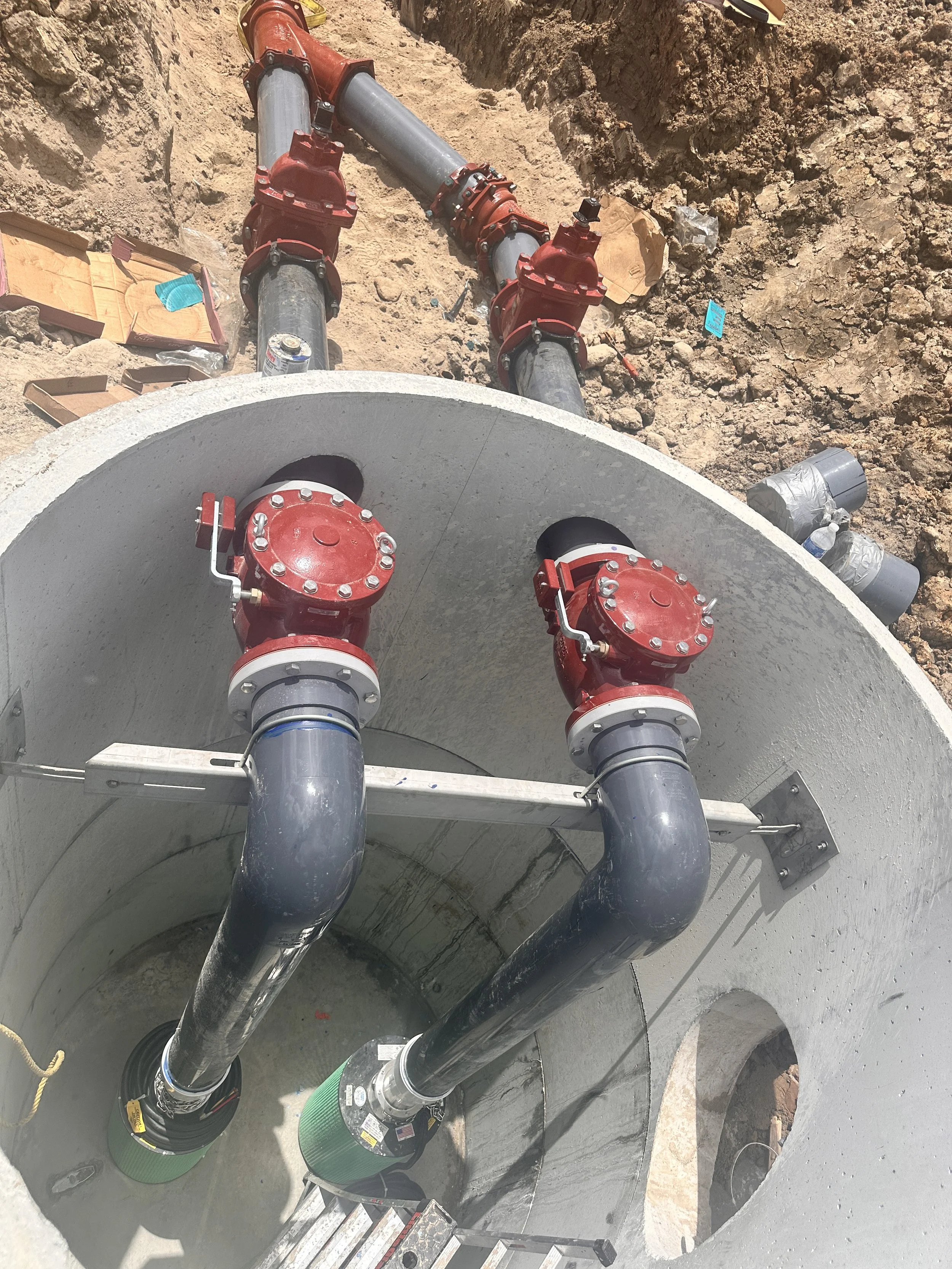 Close-up of large underground piping system with red valves, black pipes, and a concrete tank, set in a construction site with dirt and rocky surroundings.