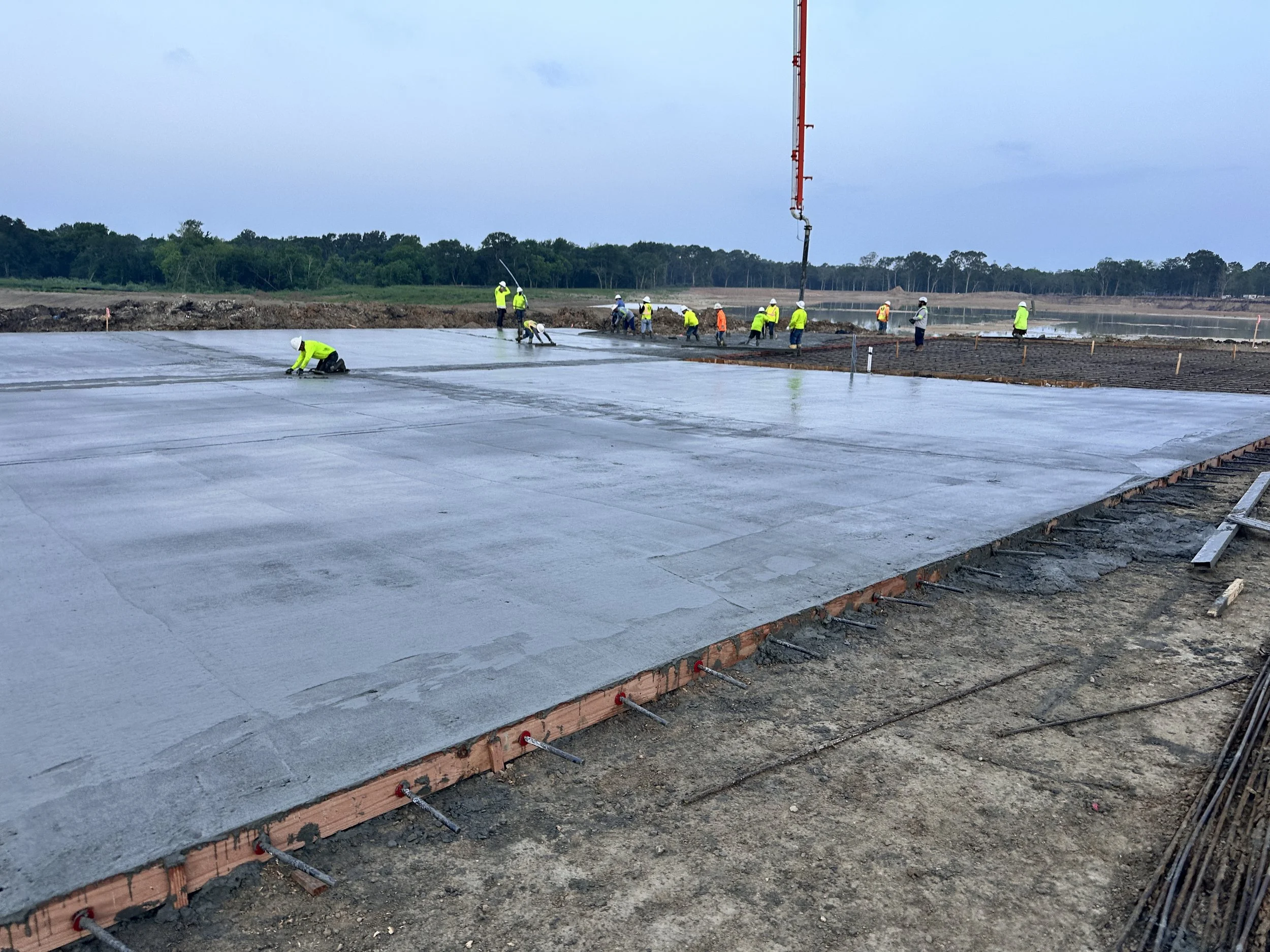 Construction workers in high-visibility clothing and helmets pouring concrete on a large foundation at a construction site during daytime.