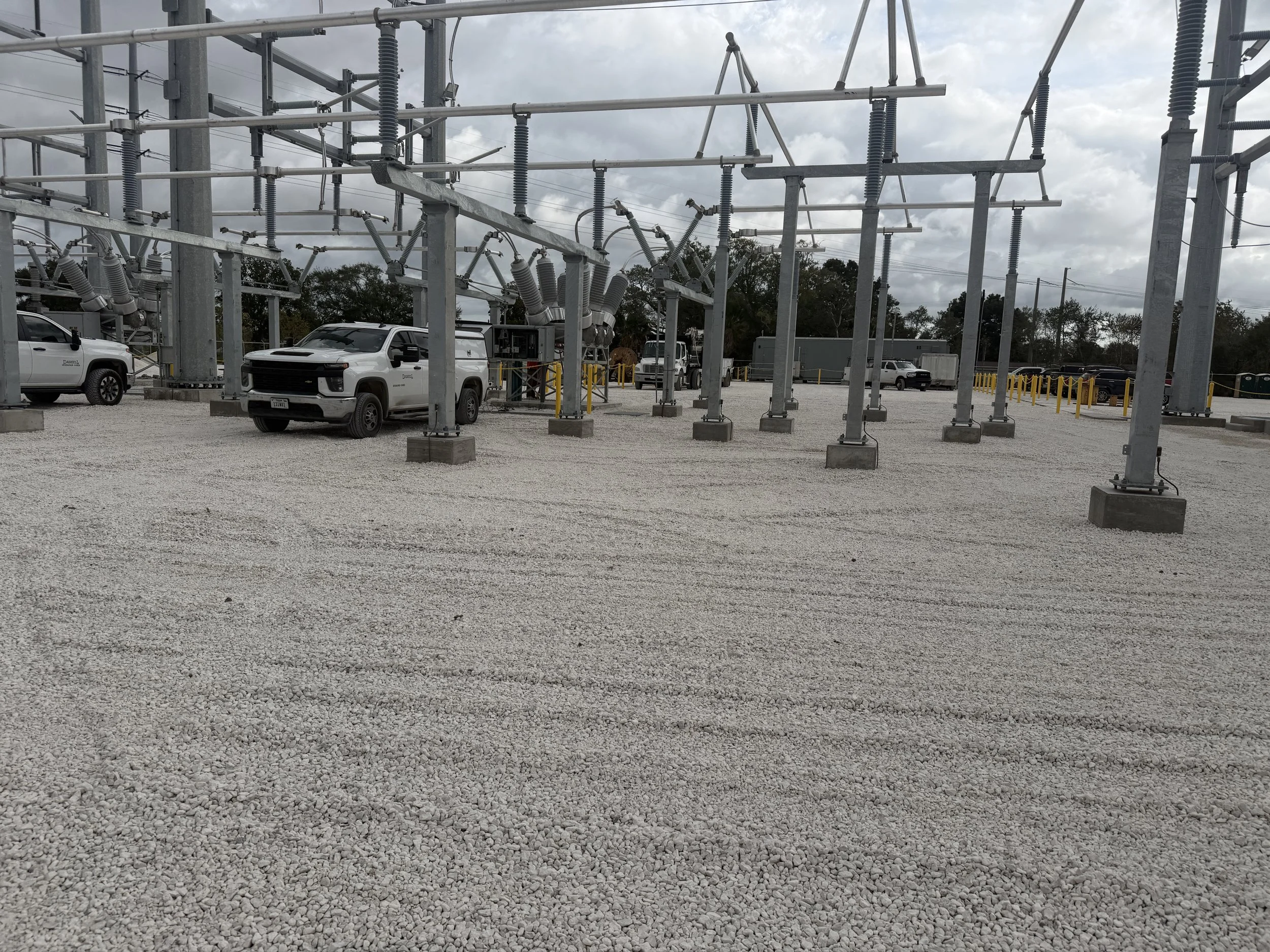An electrical substation with metal framework, transformers, and support structures, parked with several white and gray trucks on a gravel lot under a cloudy sky.