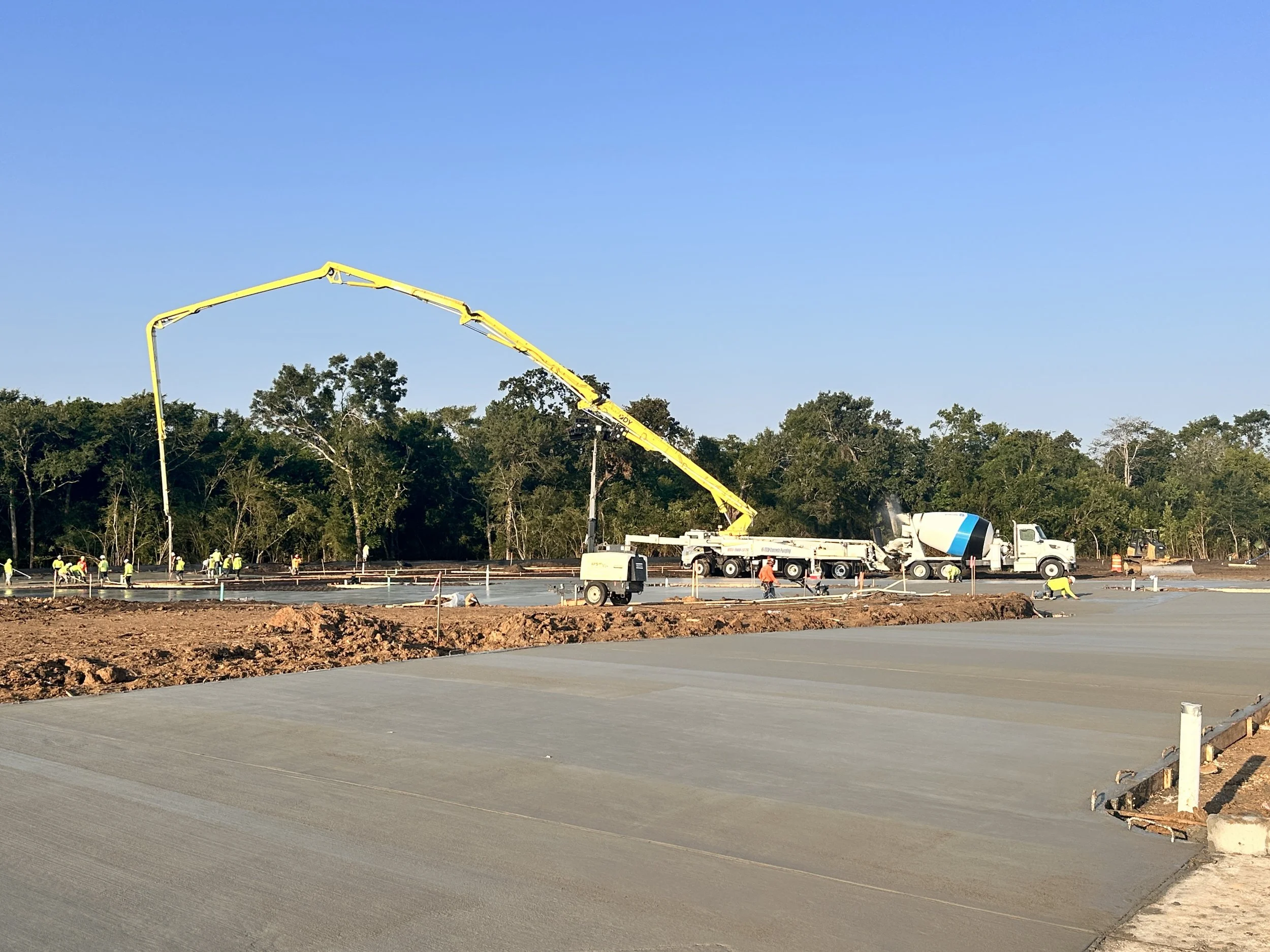 Construction workers are pouring and finishing concrete on a new roadway with a cement mixer truck and concrete pump truck on site. Trees are in the background.