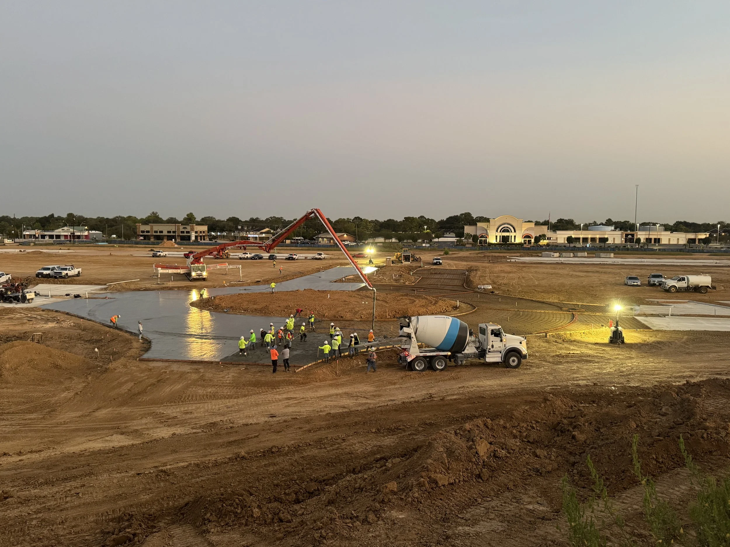 Construction site with workers pouring concrete for a building foundation, a concrete mixer truck, and construction vehicles at dusk.