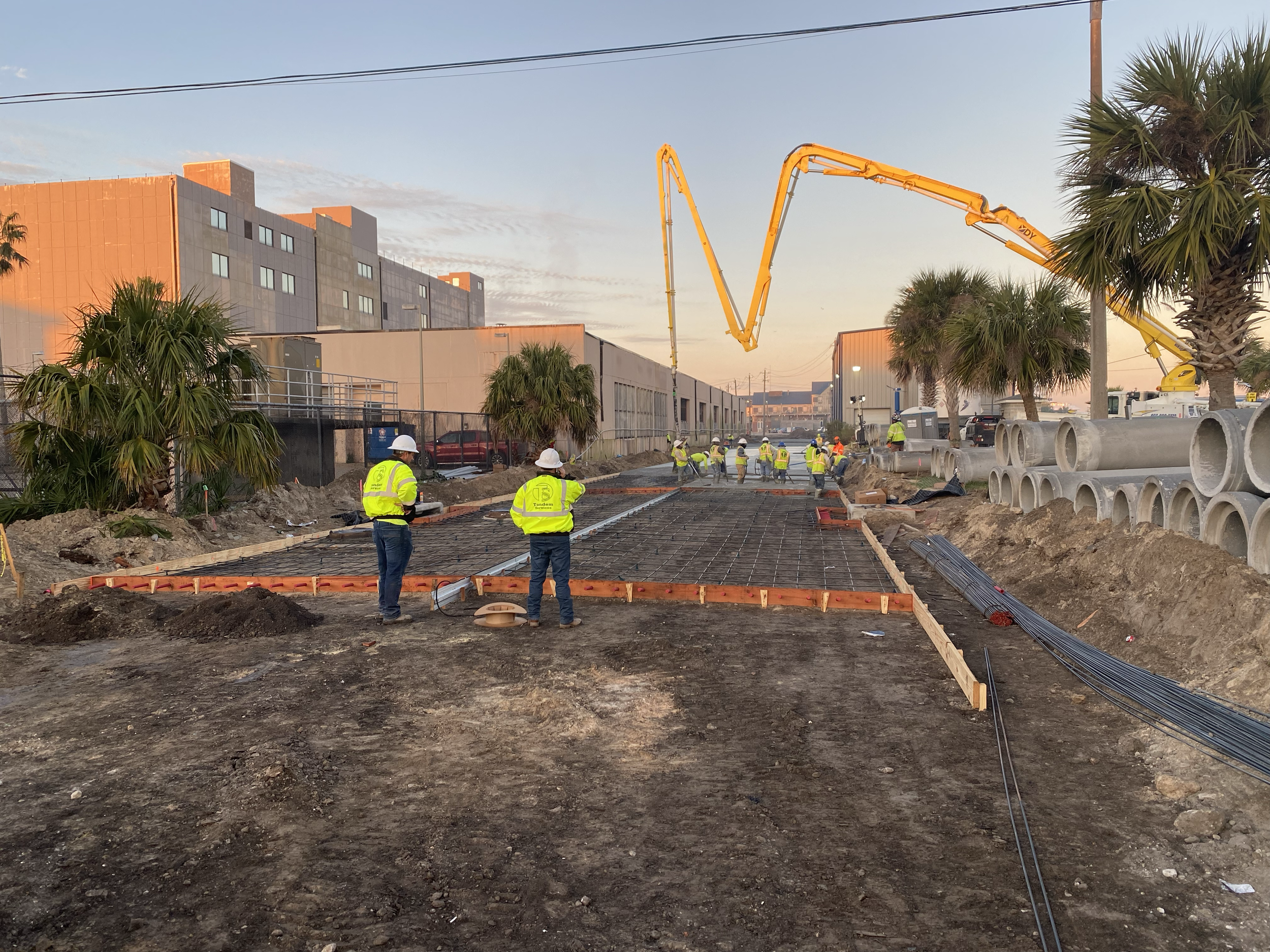 Construction site with workers wearing safety vests and helmets, pouring concrete for a walkway, with large concrete pipes stacked nearby, and a concrete pump truck in the background, during sunset