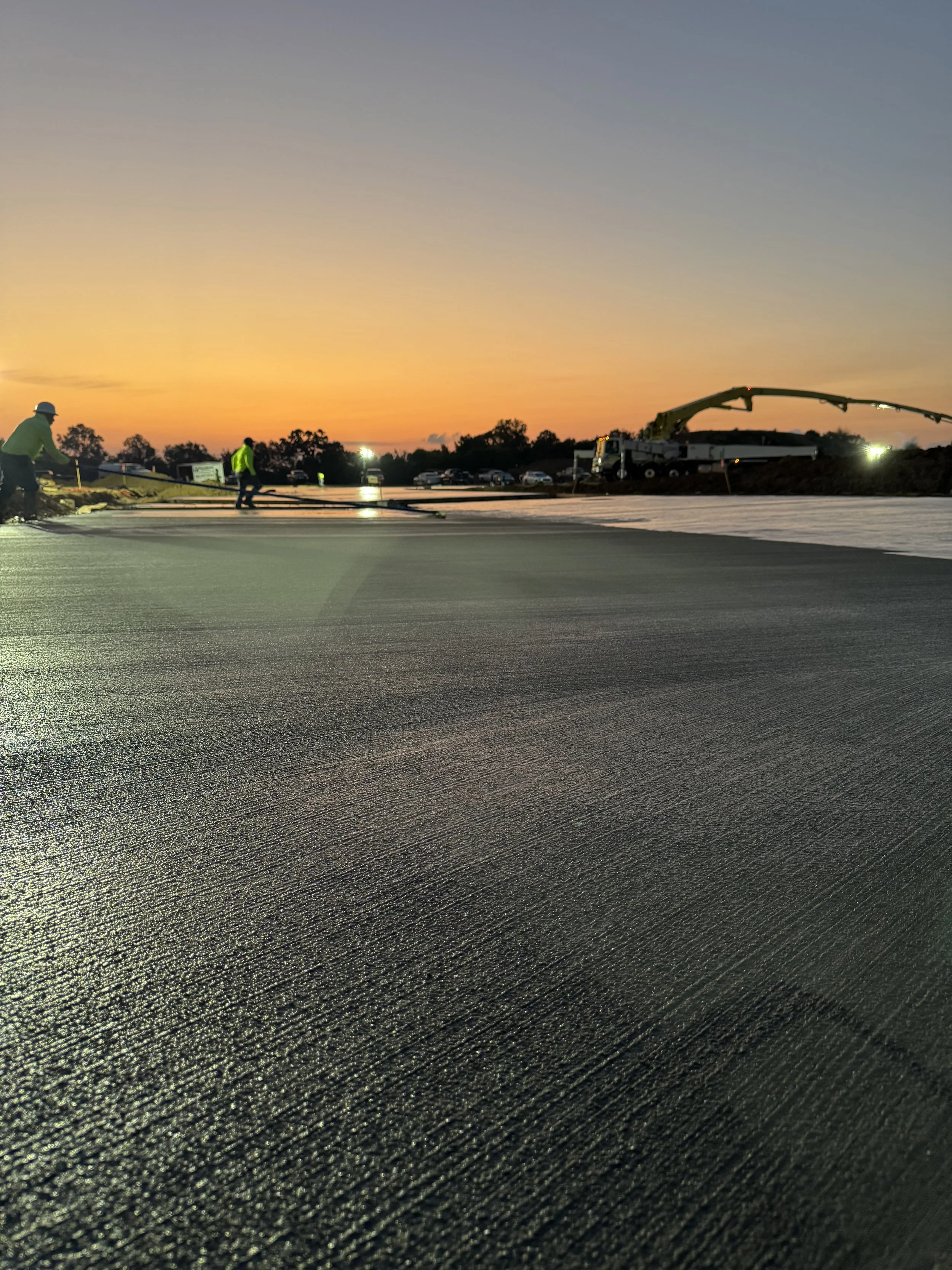Workers laying new asphalt on a road at sunset with construction trucks in the background.
