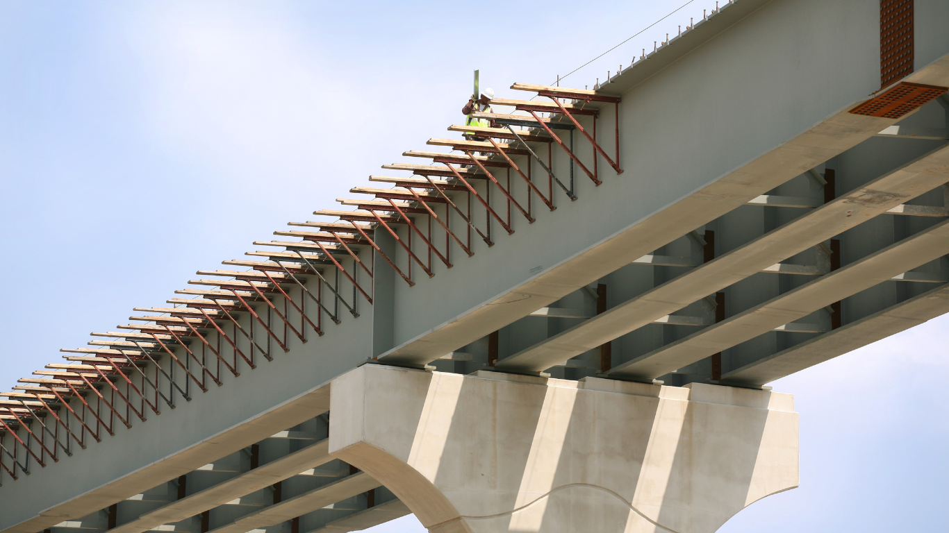 Close-up of a construction worker in a safety vest and helmet working on the underside of a bridge, which is supported by large concrete and steel beams.