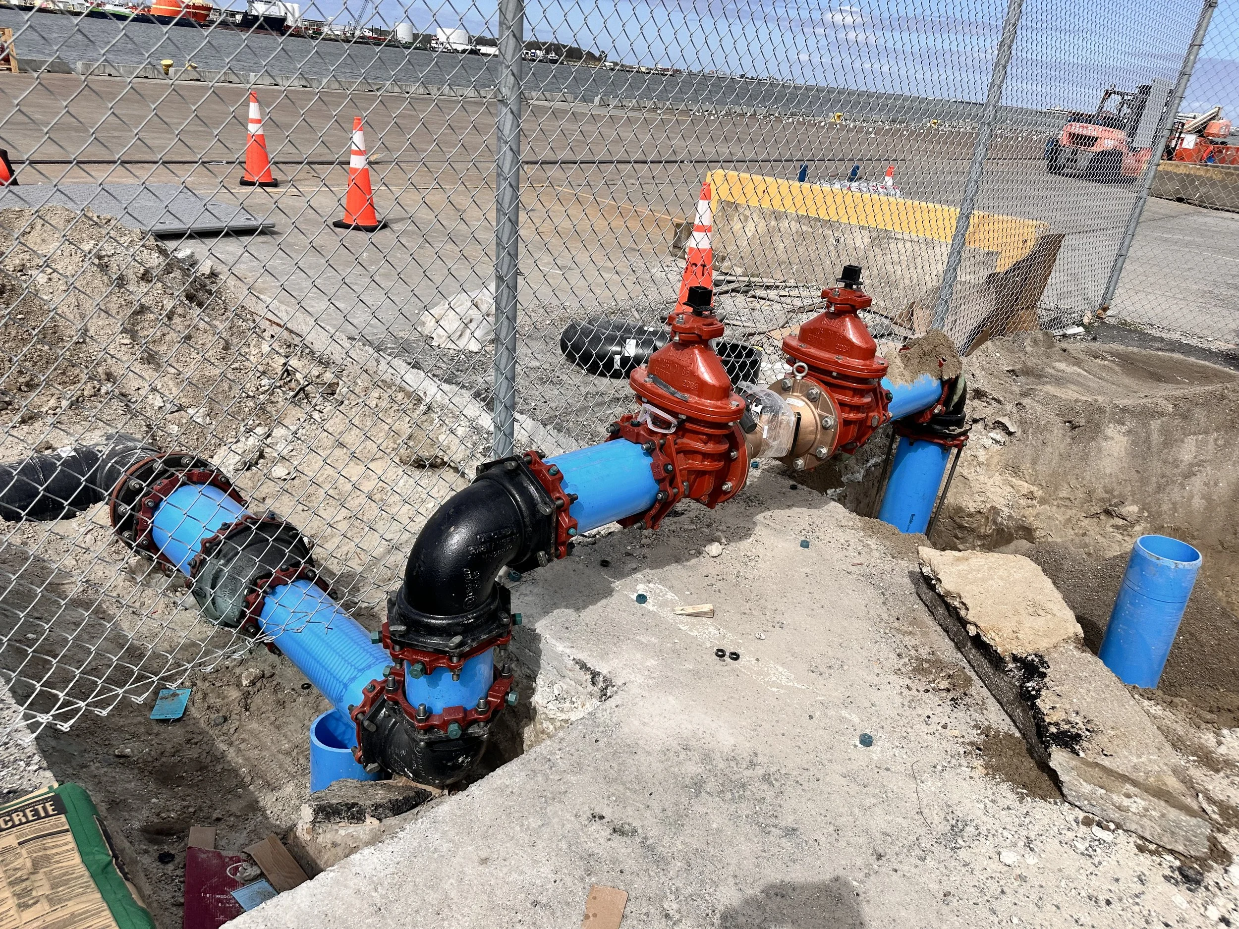 Construction site with plumbing pipes and valves, orange safety cones, chain-link fence, and construction equipment in the background.