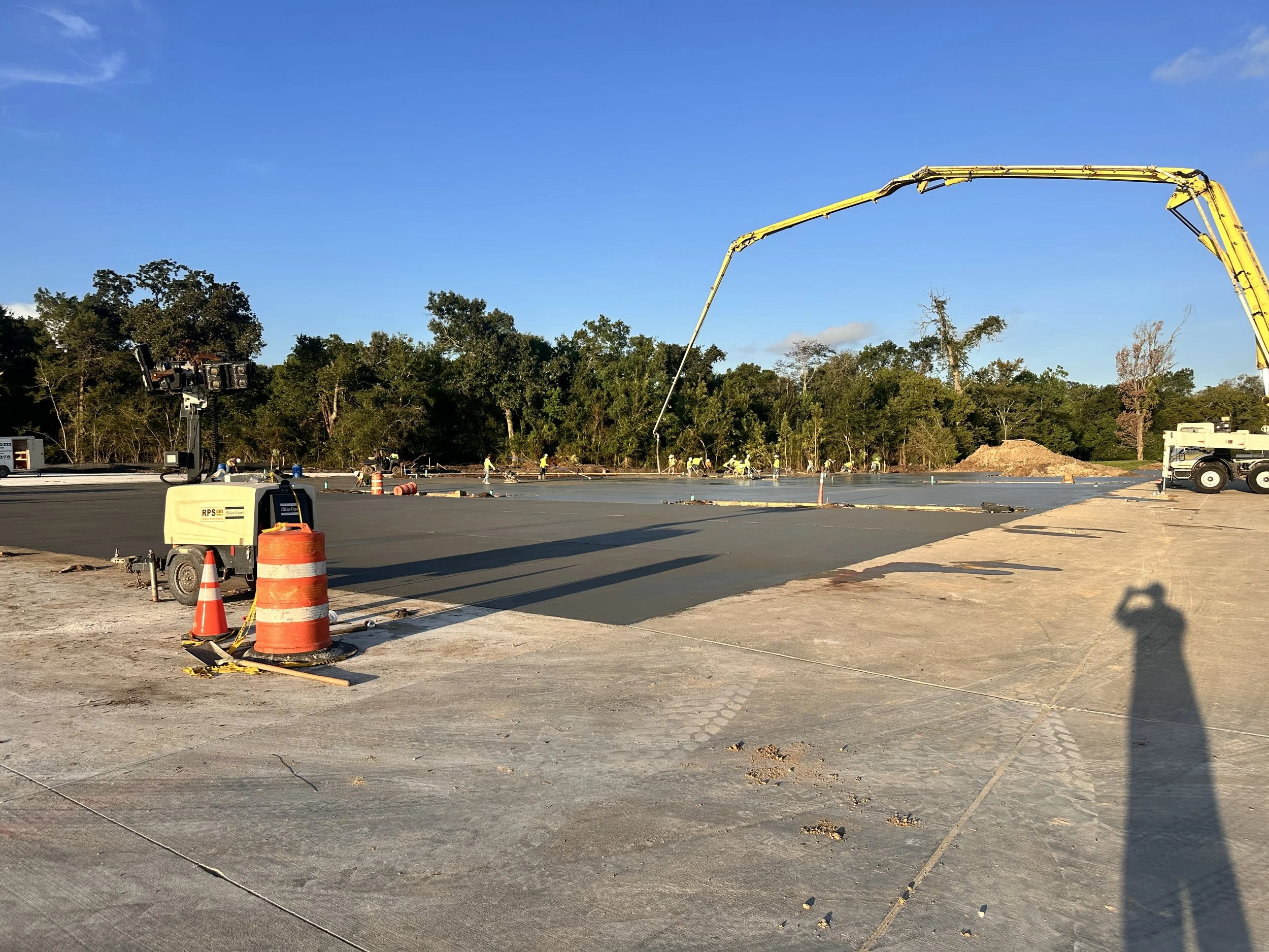 Construction site with a large concrete pour, construction workers, and heavy equipment, with trees and blue sky in the background.