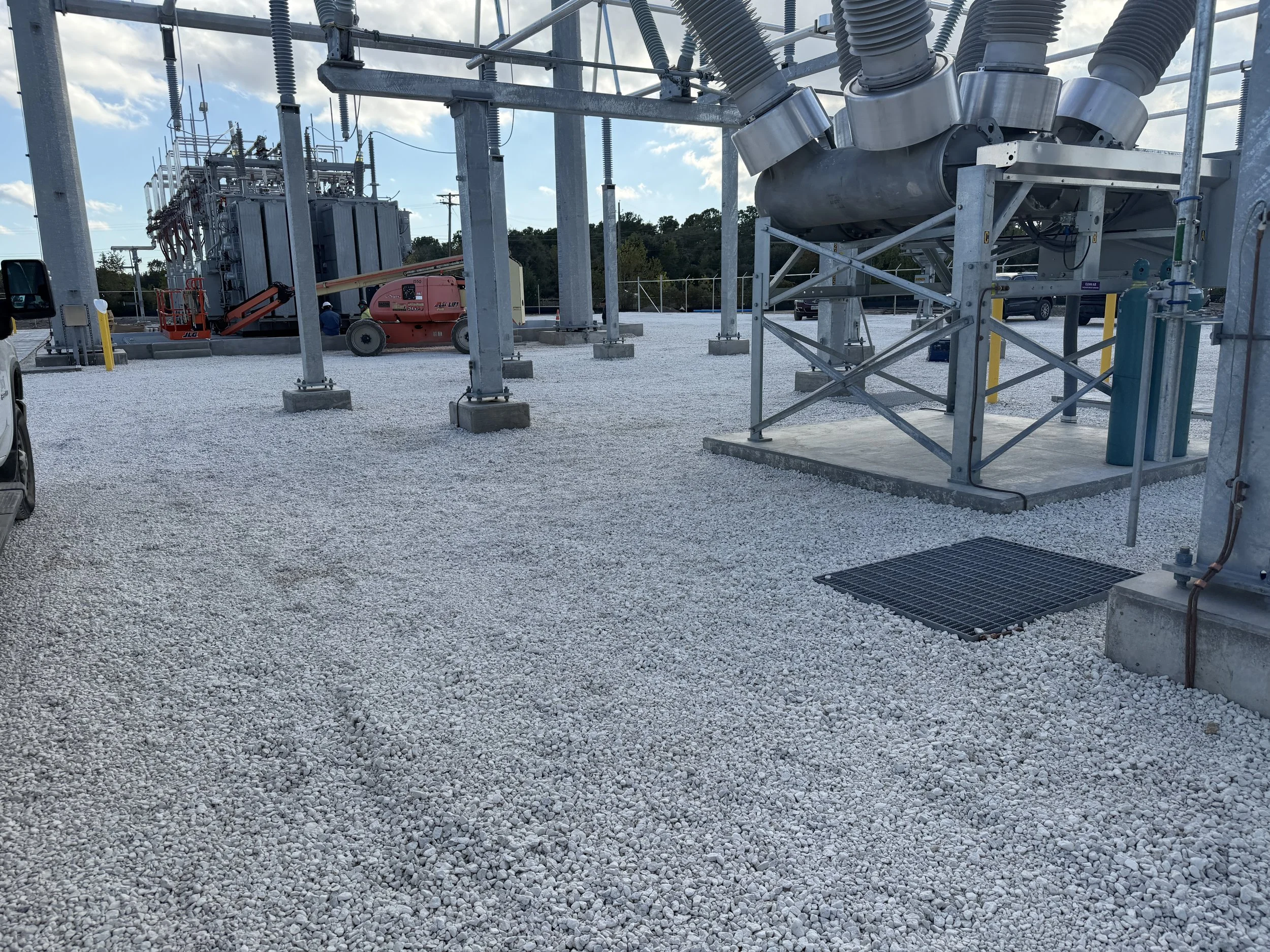 Industrial electrical substation with large transformers, metal structures, and electrical equipment, set on a gravel surface under a partly cloudy sky.