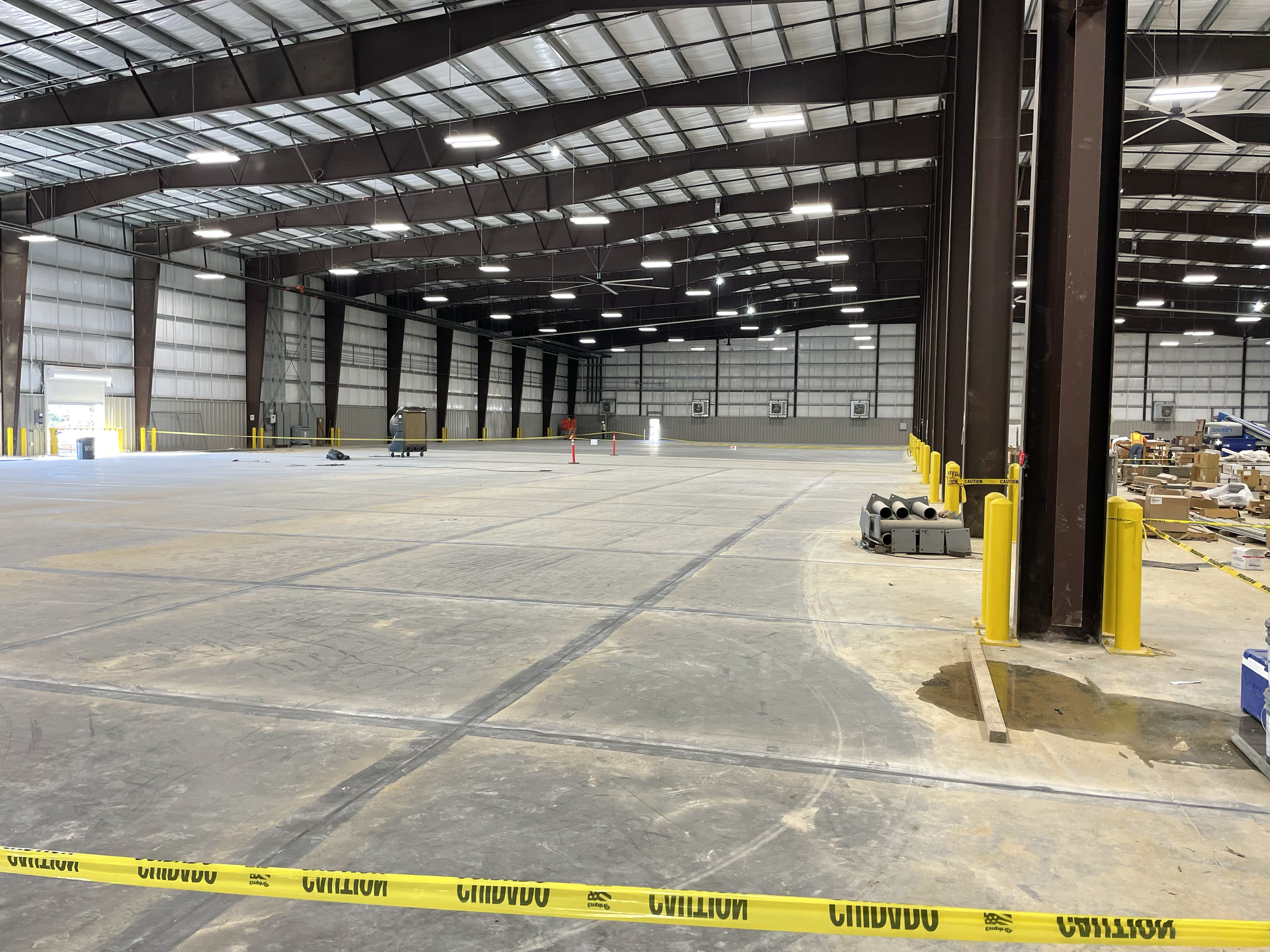Empty warehouse with yellow caution tape, some scattered equipment, and boxes along the right side. The space has a high metal ceiling with bright overhead lights.
