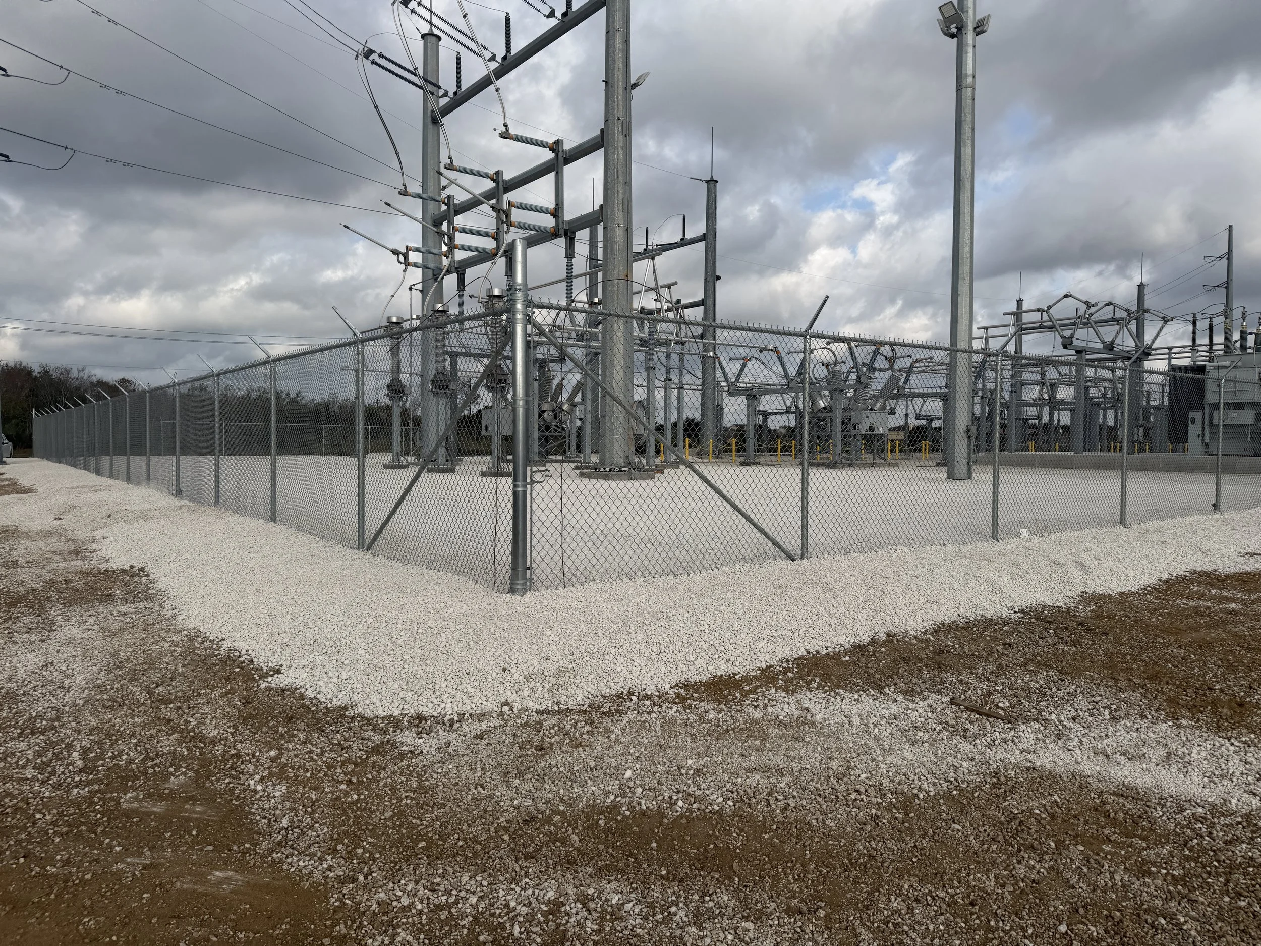 A fenced electrical substation with power transformers, insulators, and high-voltage power lines, under cloudy sky.
