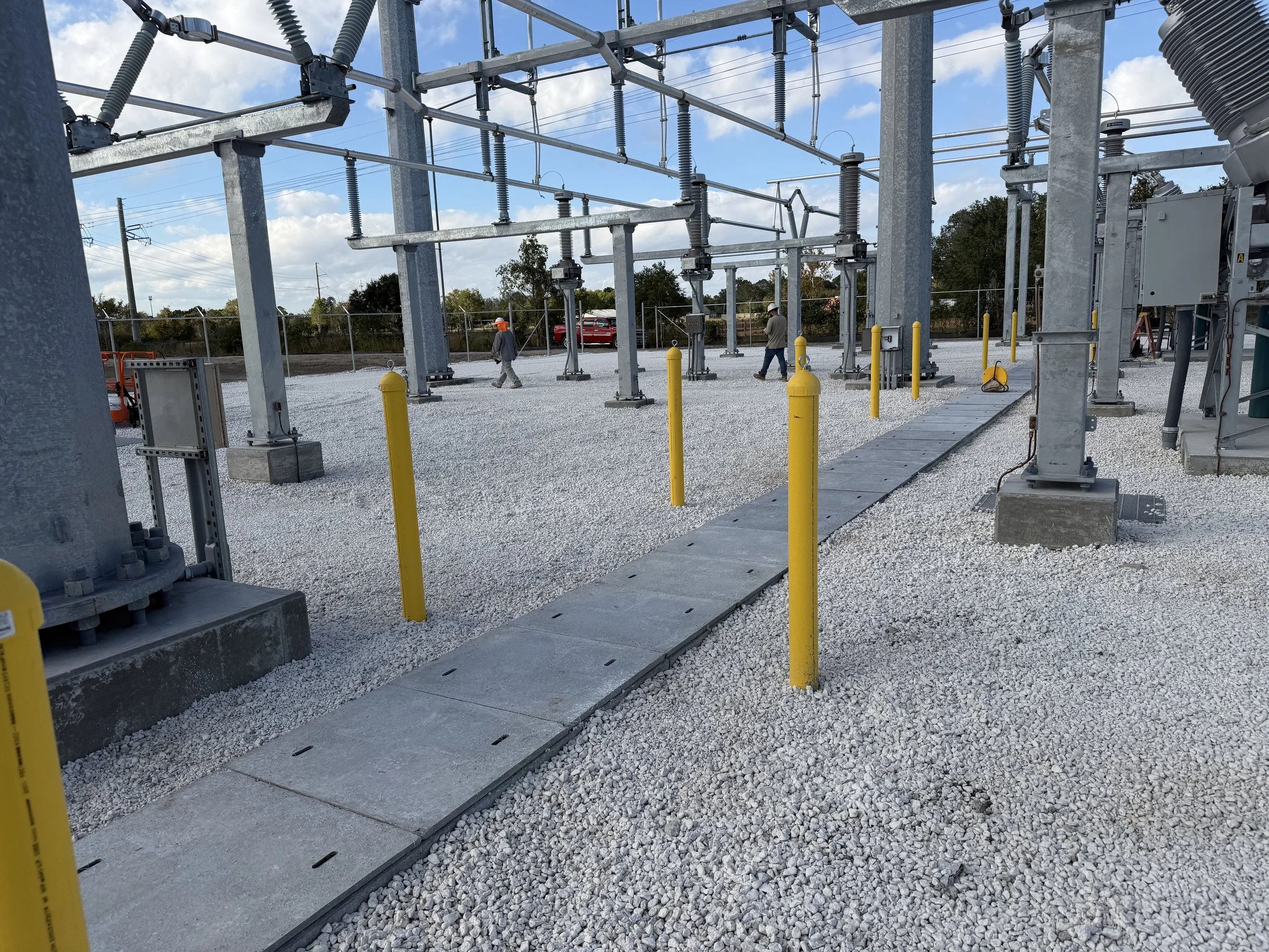 Electrical substation with metal structures, yellow bollards, a concrete walkway, and three workers, with a fence and trees in the background.