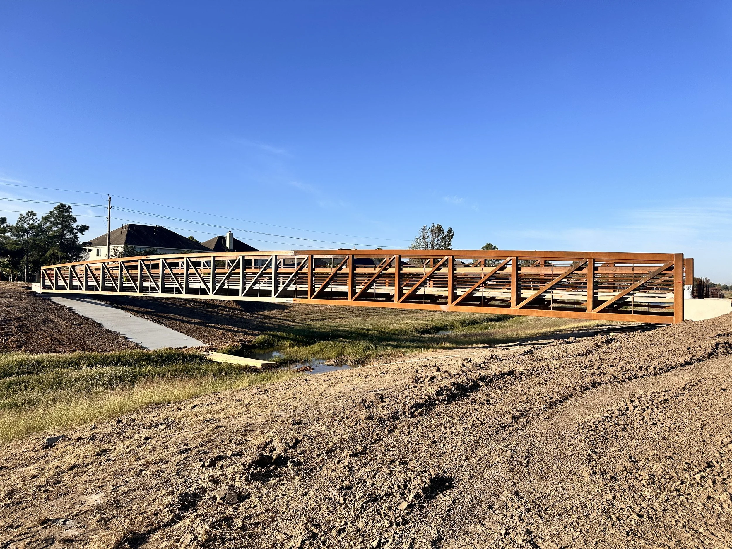 Construction site with a new wooden walkway bridge over a small pond or creek with cleared land and housings in the background under a clear blue sky.