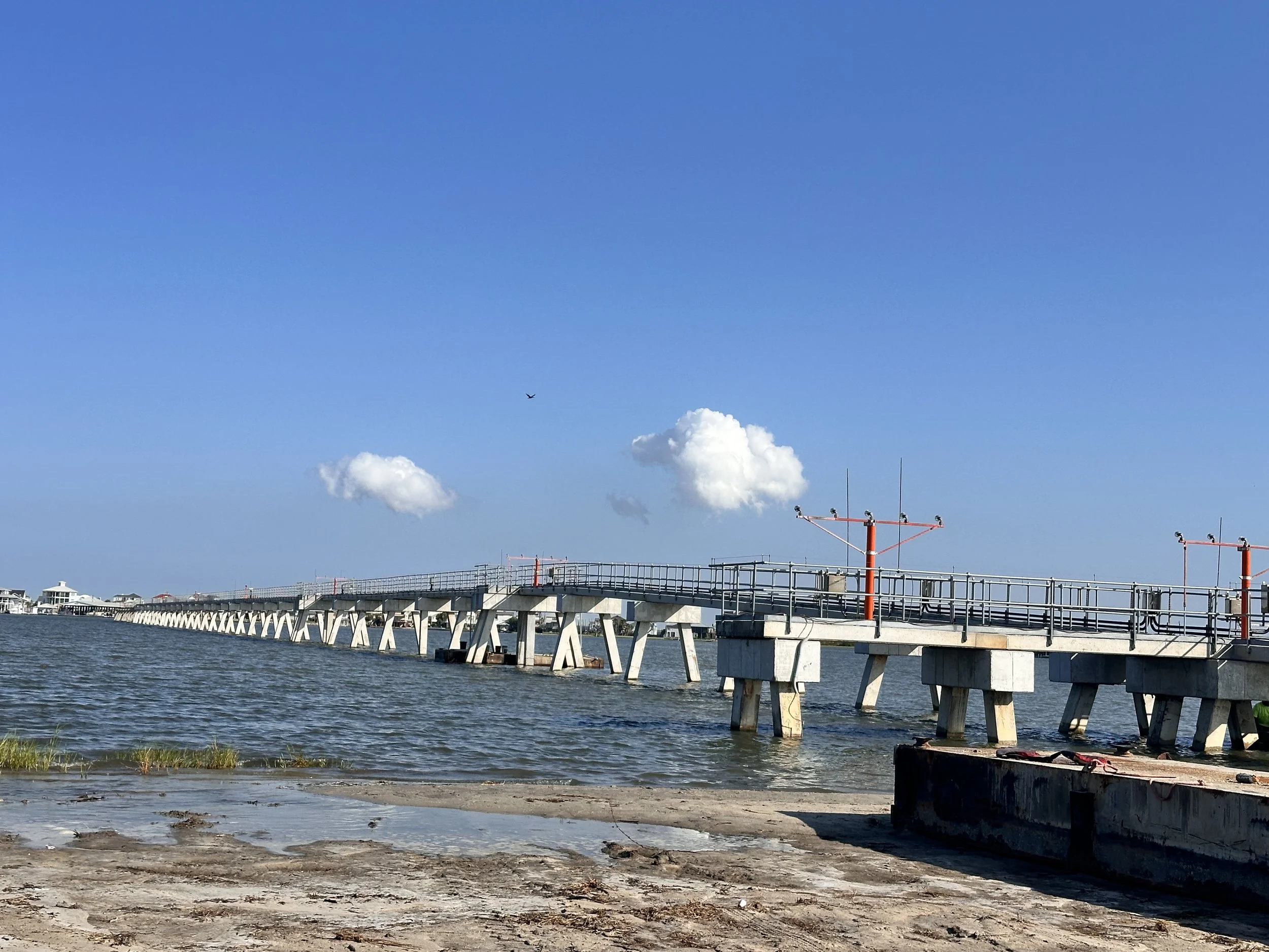 A long concrete pier extends into a body of water, with red railing and electrical poles, against a blue sky with clouds.