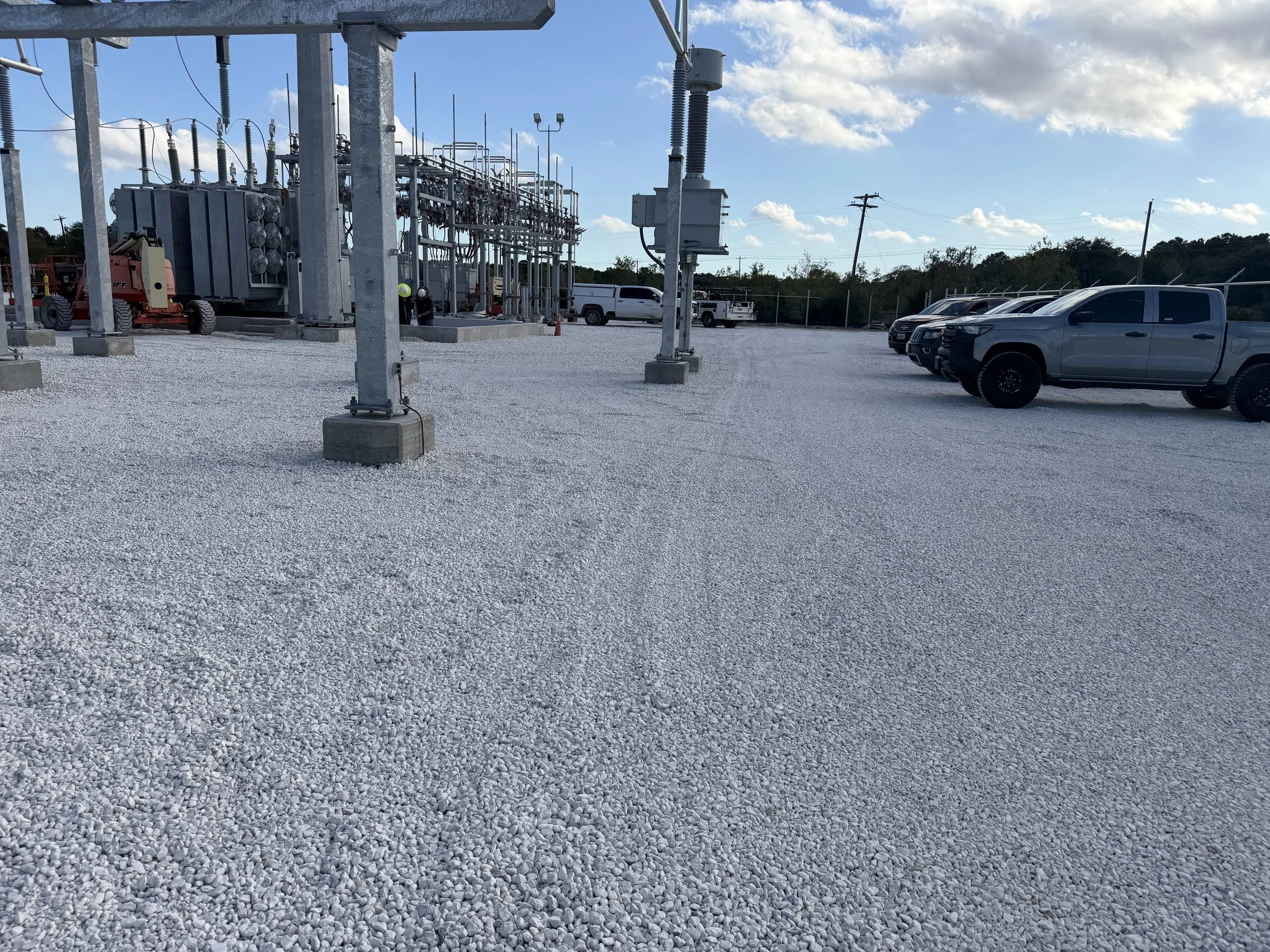 An industrial electrical substation with metal equipment and wiring, surrounded by gravel, with parked pickup trucks to the right and a blue sky with clouds overhead.