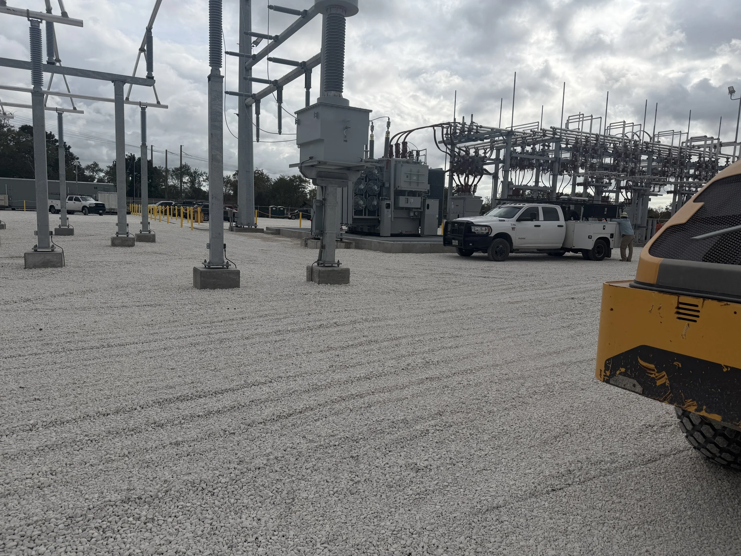 Electrical substation with equipment, vehicles, and a worker, under a cloudy sky.
