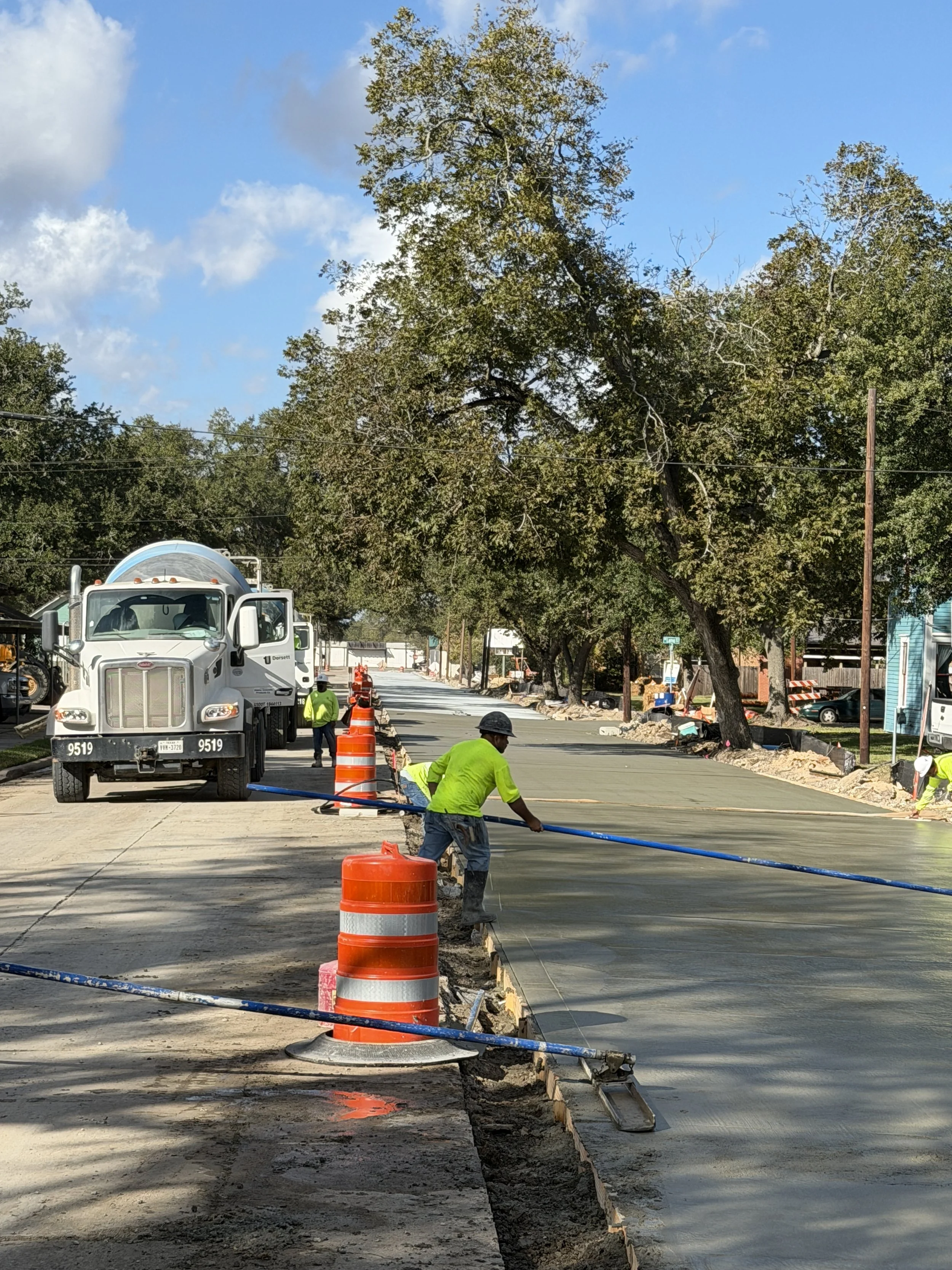 Construction workers are paving a new road, with a cement truck and orange safety cones present, under a partly cloudy sky with trees lining the street.